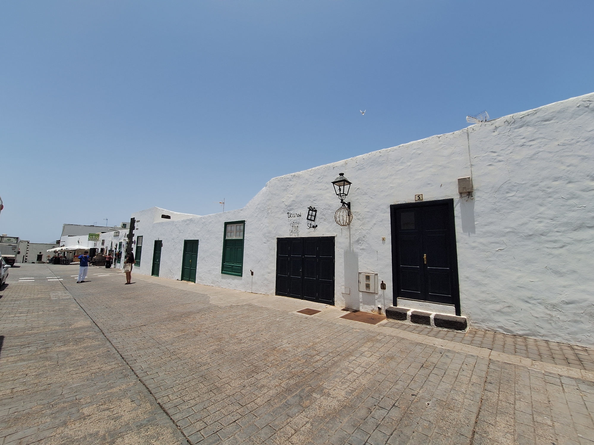 The image captures a street scene in Teguise, Spain, under a clear, bright blue sky, suggesting it is daytime with fair weather. The foreground is dominated by a cobblestone street and sidewalk. On the right side of the street, a white building with a textured facade is prominently featured. This building has several dark-colored doors and windows, including a large double door. A decorative black lamppost is mounted on the wall of the building. Some graffiti or decorative text is visible on the wall, appearing to say "Ticarol 1997 Shop".
Further down the street, to the left, more white buildings are visible, also with doors and windows. Several people are walking on the street and sidewalk. One person in the middle distance, wearing a blue shirt and white pants, appears to be holding a phone or camera. Another person, a woman in a light-colored dress, is standing further down the street. There are also market stalls set up on the left side of the street, with people browsing. A vehicle is partially visible on the far left. The overall impression is of a lively street scene in a historical town on a sunny day. A small bird is visible flying in the sky.