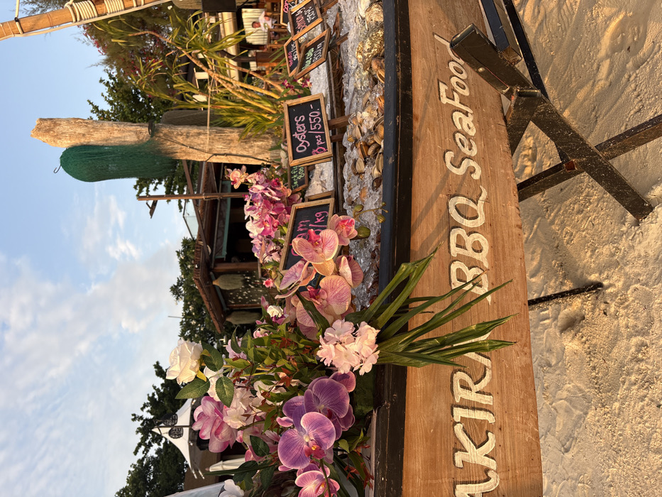 An outdoor seafood display is positioned on a sandy beach in Ban Ko Li Pe, Thailand, illuminated by warm late afternoon or early evening sunlight. The primary subject is a weathered wooden boat, serving as a display stand, supported by dark metal cross-stands. The boat's side features carved and white-painted text reading "AKIRA BBQ SeaFood."
Within the boat, various seafood items are arrayed on a bed of ice. Small, freestanding chalkboards indicate offerings and prices, including "Oysters 3 pcs / 550.-," "Jackfish 800," and "Red Snapper 600 kg." The boat is decoratively adorned with a large arrangement of artificial flowers, primarily pink and purple orchids, interspersed with white roses and green foliage.
In the mid-ground, behind the seafood display, several structures characteristic of a beachside resort or restaurant are visible. To the left, a structure features a white conical roof. Centrally, a large, rough-hewn wooden post stands, draped with a dark green fishing net-like material, with a light fixture attached to an adjacent pole. Further back, a rustic wooden building with a thatched or wooden roof is present. Tropical vegetation, including palm trees, is distributed throughout the background. A single, blurry individual is discernible seated at a table under a thatched roof structure in the far right background. The sky overhead is clear blue with scattered white clouds.