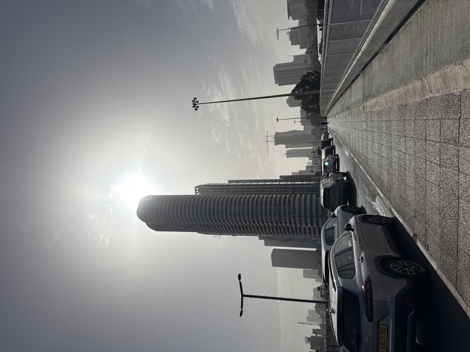 The image depicts an urban street scene in Tel Aviv-Yafo, Israel, observed from a bridge or overpass. The view is oriented towards the east or southeast, with the sun positioned directly behind the prominent Arlozorov Towers, creating a strong backlight and haze across the upper sky.
The environment includes a multi-lane asphalt road on the left, congested with numerous passenger vehicles. The closest vehicle, a silver Toyota Corolla Cross SUV, bears an Israeli license plate "430-11-704". Other vehicles, including several SUVs and sedans, extend in a queue further down the road, indicating a traffic jam. A green light, possibly a traffic signal or vehicle reflection, is visible above the third vehicle in line.
To the right of the road, a wide pedestrian and bicycle path is paved with square grey tiles, separated by a curb. The path appears reflective, possibly due to dampness. A grey metal safety railing lines the far right edge of the path. One person is faintly discernible walking on the path in the distance, silhouetted against the bright background.
Dominating the skyline is the distinctive Arlozorov Towers, a multi-story, twin-structure skyscraper with curved, segmented facades. Numerous other high-rise buildings, some complete and others under construction with visible cranes, form a dense urban backdrop extending into the hazy distance. Two streetlights are present: one on the left with a single rectangular fixture, and a taller one on the right with four square-arranged fixtures. The sky is bright but hazy with minimal cloud cover.