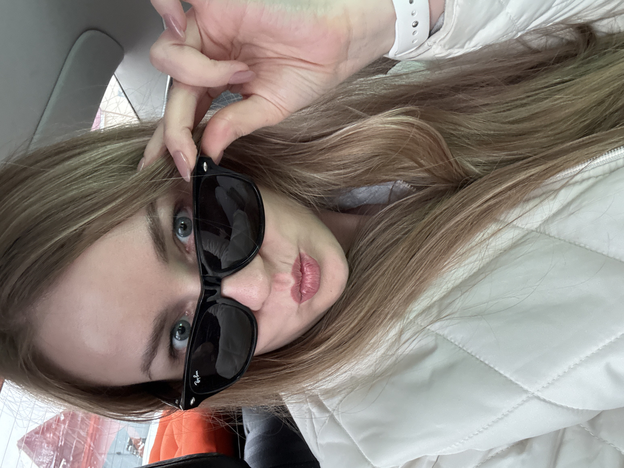 The image shows a young woman in a car, holding Ray-Ban sunglasses. She is wearing a white, quilted jacket and a white Apple watch. The location appears to be Chaikovsky, Russia, judging by exterior reflections of Cyrillic text on building signs, partially visible. The woman is looking at the camera with a slightly puckered expression. The interior of the car, including the window and headliner, is visible. The image is taken from a slightly downward angle.