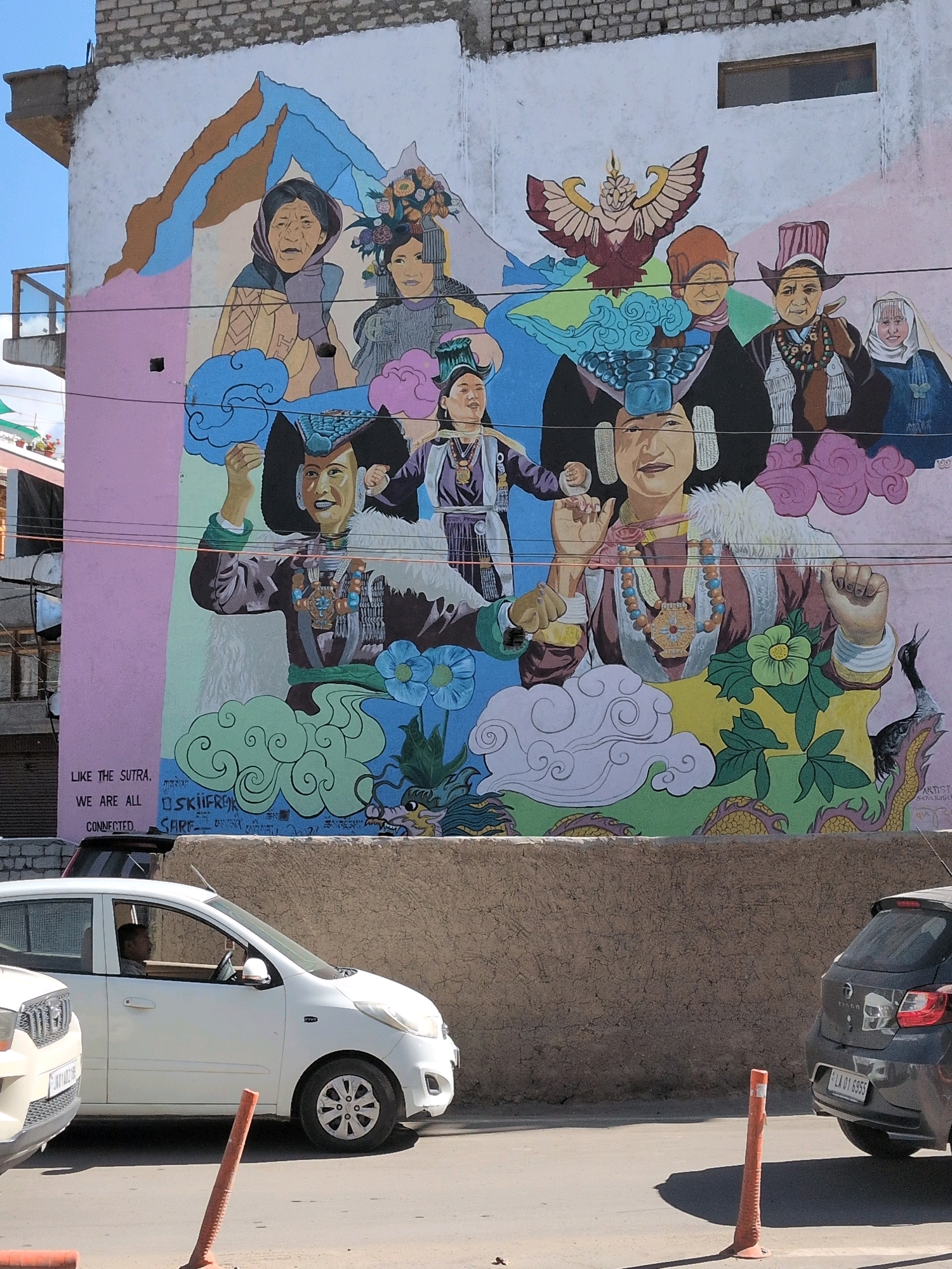 This is a photograph of a mural on the side of a building in Leh, India. The mural depicts a group of people with traditional clothing and headdresses, along with various symbolic elements such as mountains, clouds, a bird, and a dragon. There is text on the lower left side of the mural that reads "LIKE THE SUTRA. WE ARE ALL CONNECTED." Below the mural, a white car is parked on the street, with a person driving it. A dark-colored car is parked behind it. The scene appears to be outdoors, with a blue sky visible in the upper left corner. The building has a brick facade at the top. The street is paved, and there are traffic cones present. The overall impression is a vibrant and culturally rich scene.