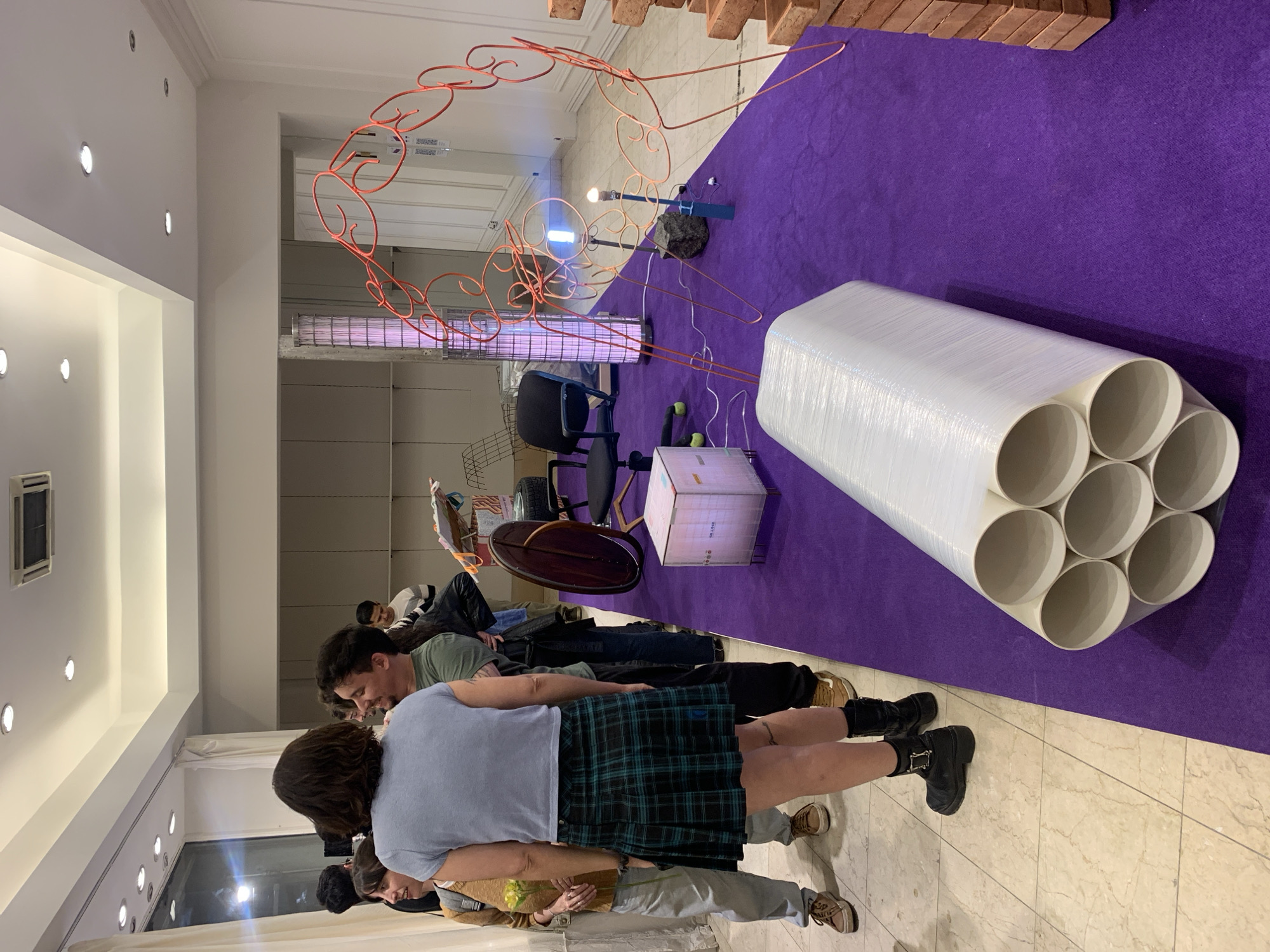 The image shows an art installation in Buenos Aires, Argentina. The installation is set on a purple carpet and includes a stack of bricks, a red wire sculpture, a box of glowing tubes, a rock with an illuminated bulb, and a large, white, multi-tubed object wrapped in plastic. Several people are standing and looking at the art. The location is an indoor space with bright lighting.