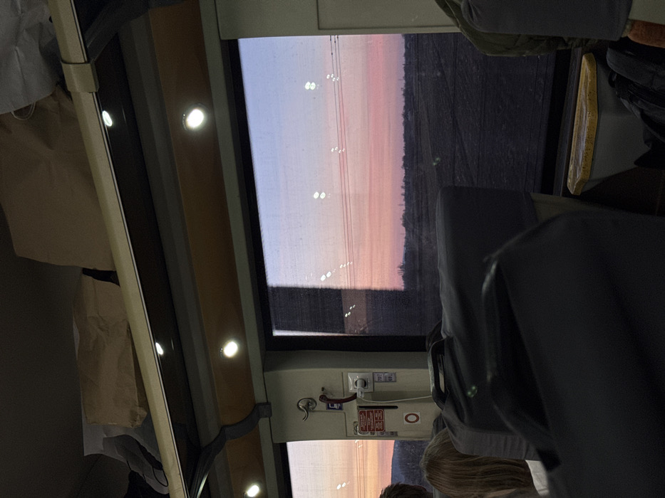 The image captures the interior of a passenger train car within the city of Ungheni, Romania, at either sunrise or sunset, viewed from a seated perspective looking towards the windows. The exterior view reveals a gradient sky transitioning from light blue to vibrant pink and orange, above a flat, dark landscape consistent with fields or undeveloped land. Multiple horizontal power lines are visible across the sky, reflecting interior train lights. Inside the train, recessed ceiling lights illuminate a beige overhead luggage rack containing several light brown paper bags and a light-colored shoe. A wall-mounted utility panel between the windows features a hook, a red emergency pull handle, and an electrical outlet, with a label displaying "20" and "50". In the foreground, dark fabric-covered seatbacks are prominent, with a dark grey luggage item resting on one seat. The back of a person's head with blonde hair is visible on the far left. A yellow-rimmed waste receptacle is positioned beneath the right window.