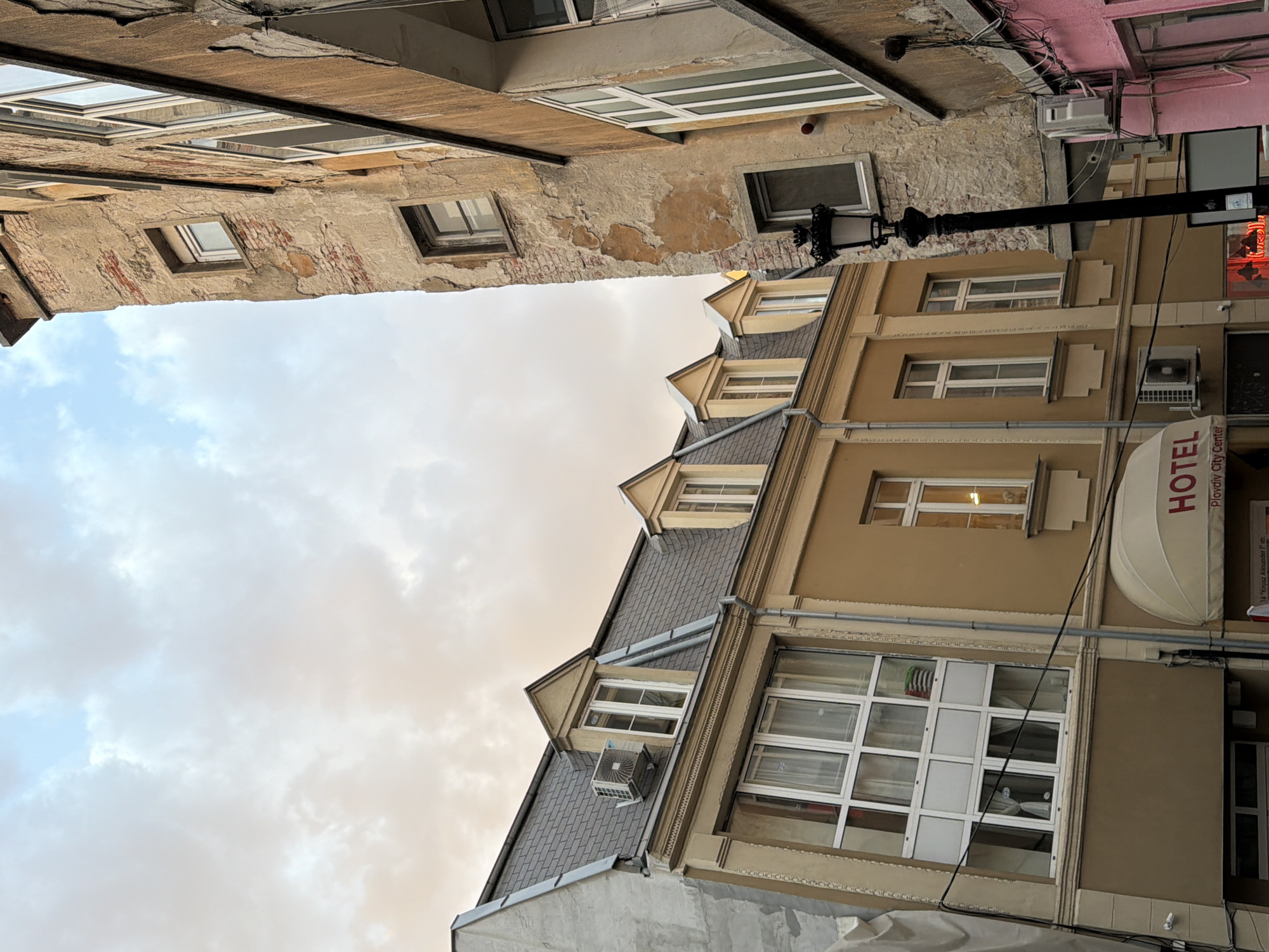 The image shows a street scene in Plovdiv, Bulgaria, looking upwards between buildings. On the right is a building with a "HOTEL Plovdiv City Center" awning. The building is multiple stories with tan facade, white windows and architectural details. It has a gray roof with dormer windows. An air conditioning unit is mounted on the dormer roof. The building on the left is older, with peeling plaster exposing brick beneath. It also has windows, some with white frames. Above, the sky is visible, with clouds present. A street lamp is attached to the side of the buildings. Wires are running across the facade of the hotel building.