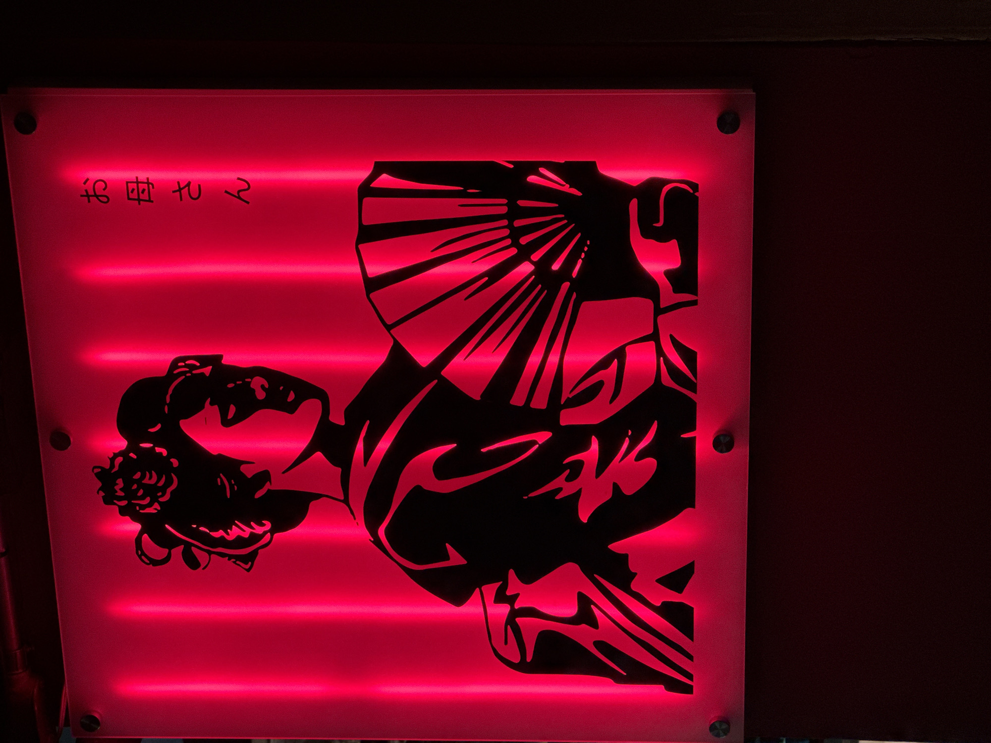 The image shows a red neon-lit sign depicting a silhouette of a Japanese geisha holding a fan. Japanese characters reading "お母さん" (Okasan), meaning "mother", are visible on the right side of the sign. The sign is mounted on a wall, and the lighting creates a vibrant, moody atmosphere. The image was taken in Bangkok, Thailand.