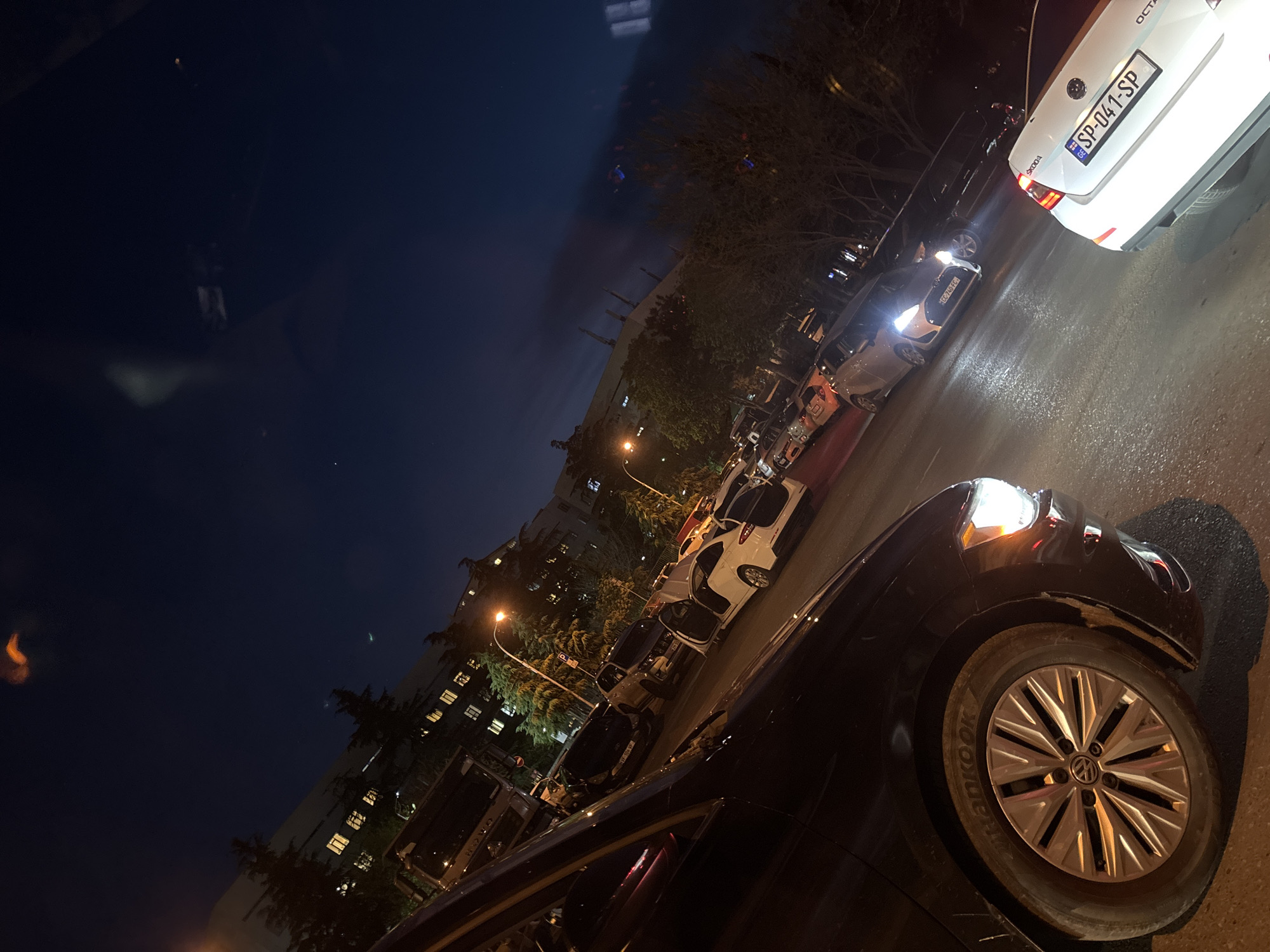 The image depicts a nighttime street scene in a residential area of Tbilisi, Georgia, with multiple cars parked along the curb. The perspective is tilted, suggesting a low-angle shot.
In the foreground, the front quarter of a dark-colored car, identifiable by its Volkswagen wheel cap and Hankook tire, is visible with its headlight illuminated. Further down the street, a white Skoda Octavia is prominently featured from the rear, displaying a Georgian license plate "SP-041-SP" along with the "SKODA" and "OCTAVIA" branding. Other parked vehicles line the street, one of which shows a partial license plate "CC-740-FC".
The background features multi-story residential buildings with illuminated windows, indicating people inside. Trees, some bare and some leafy, are interspersed along the street and around the buildings. Streetlights cast an orange-yellow glow, combining with the bright white car headlights and red taillights to illuminate the scene. The road surface appears slightly reflective, possibly from dampness. No human activity is visible, conveying a quiet urban night.