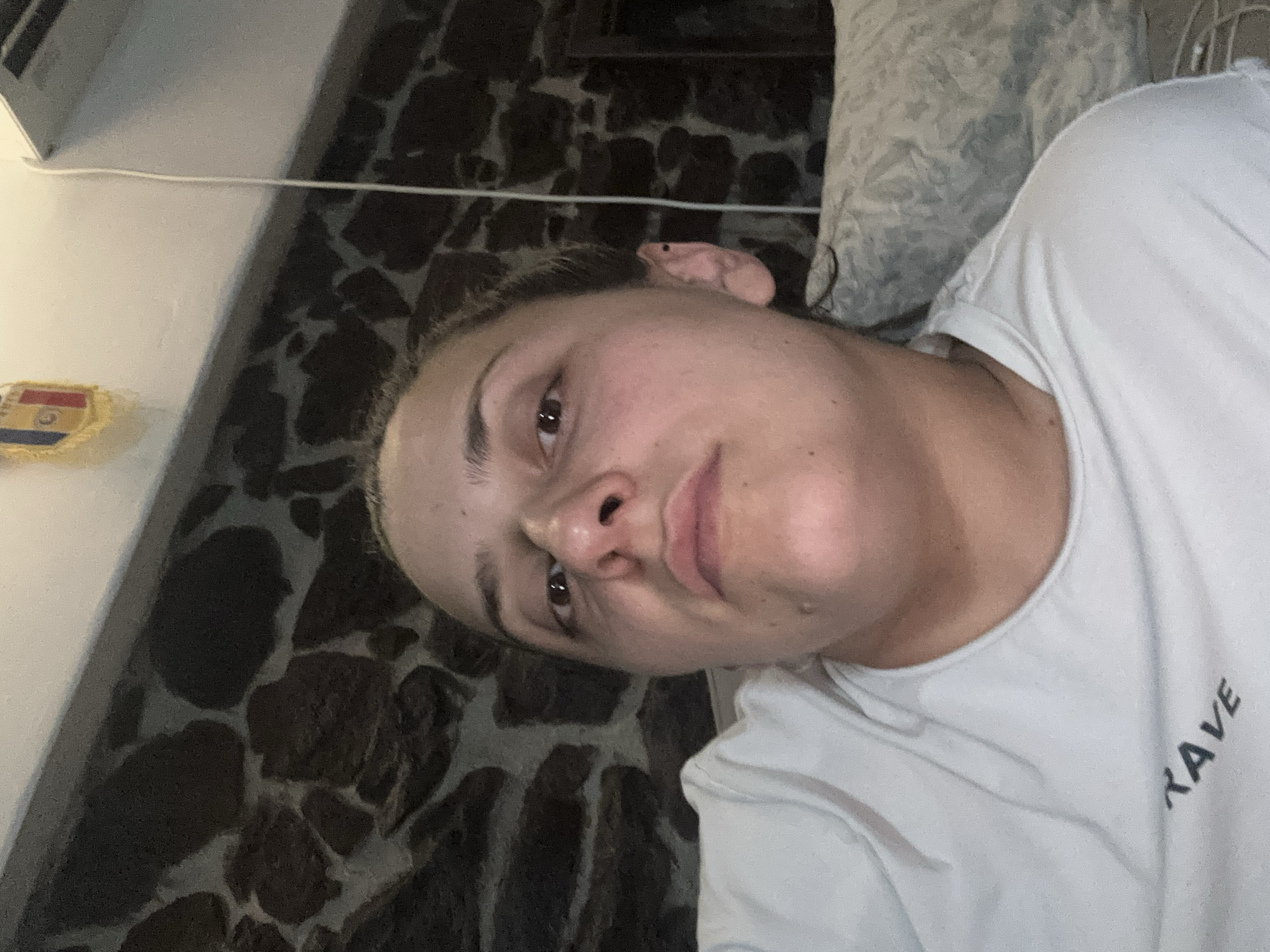 A young woman is visible from the chest up in an off-center, rotated indoor shot. She is lying against a pillow with a white t-shirt. The background features a stone wall. On the left of the wall is a wall vent and an emblem with yellow, red, and blue stripes. The location is unknown, but the request included "Belopoptsi, Bulgaria".