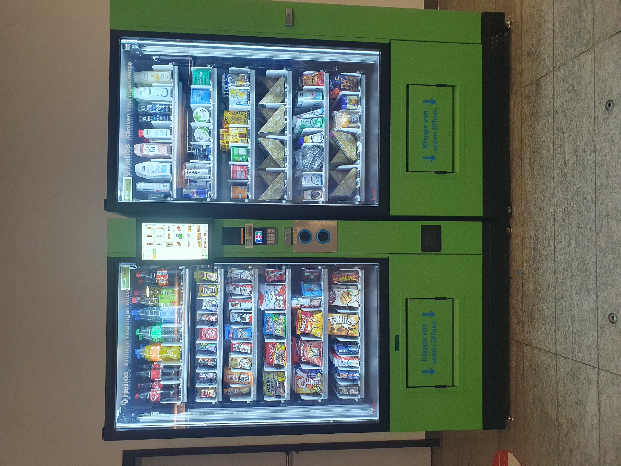 The image shows two adjacent green and black vending machines. Both machines have glass fronts displaying various products. The left machine appears to be a food and beverage vending machine, stocked with bottles of drinks and packaged snacks such as crisps, chocolates, and sweets. The right machine also contains beverages and what appear to be personal care items such as shower gels and lotions, as well as more packaged food items. Both machines have screens and keypads for selection and payment. The text on the machines and above the right machine's screen includes "Helios Kliniken Thüringen Mitte," indicating they are located at Helios Clinics in Thuringia Mitte, which is in Erfurt, Germany. Below the dispensing slots of both machines, there is a green flap with blue text that reads "Klappe von unten öffnen," meaning "Open flap from below." The machines are situated indoors on a grey tiled floor. The lighting is bright, illuminating the products within the machines.
