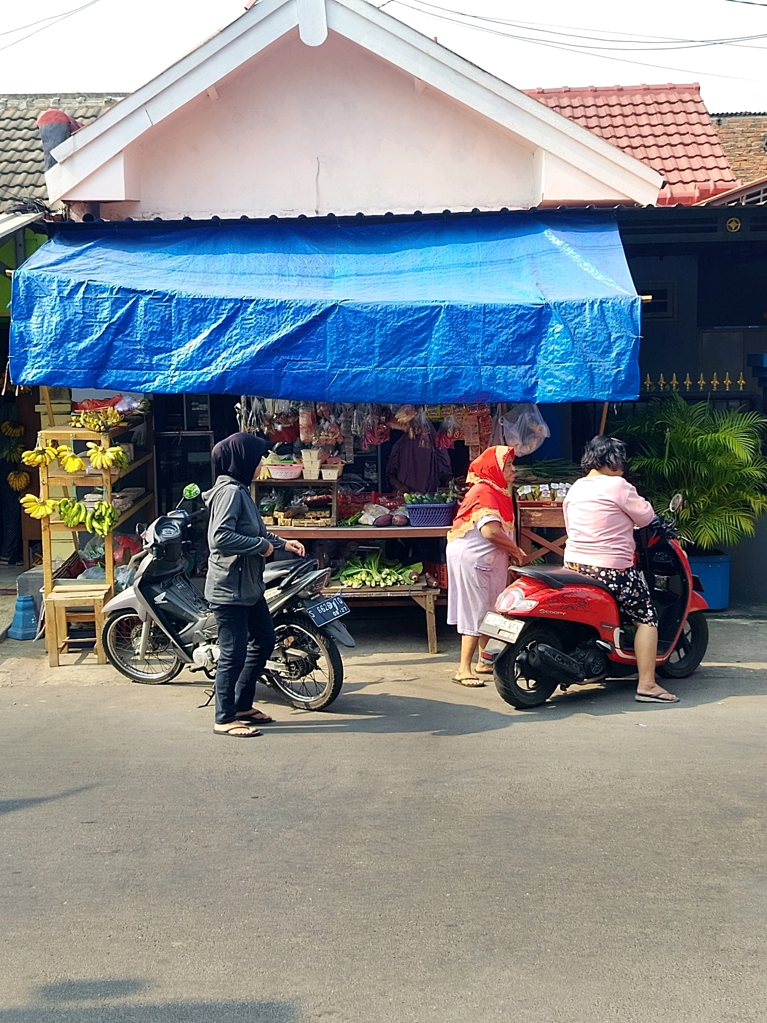 In Mojokerto, Indonesia, on a sunny day, a small roadside market is set up in front of a building with a light pink facade and a red tiled roof. A bright blue tarp provides shade over the market stalls. On the left, a woman wearing a gray hijab and jacket stands next to a silver motorcycle. The market displays various fresh produce, including bunches of yellow and green bananas on wooden shelves, and other items like vegetables and packaged goods. To the right, two women are interacting. One woman, wearing a red hijab and traditional attire, stands beside a red scooter. The other woman, on the red scooter, is wearing a pink shirt and patterned shorts. The scene captures a typical street-side commerce setting in the city.