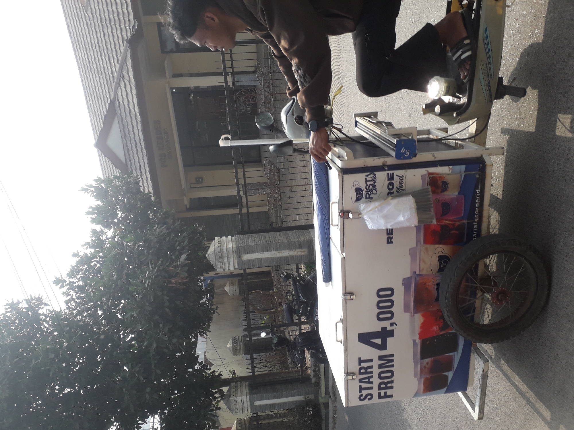 A young man is riding a scooter with a food cart attached, on a street in Cankuang, Indonesia. The man is wearing a brown hoodie and black pants, and is looking to the right. The food cart is white with a blue trim, and has a picture of drinks on the side. It also has a cooler on top. In the background, there is a building with a sign that says "POS KEBON". The street is paved, and there are trees and fences on the side of the road. The weather is sunny.