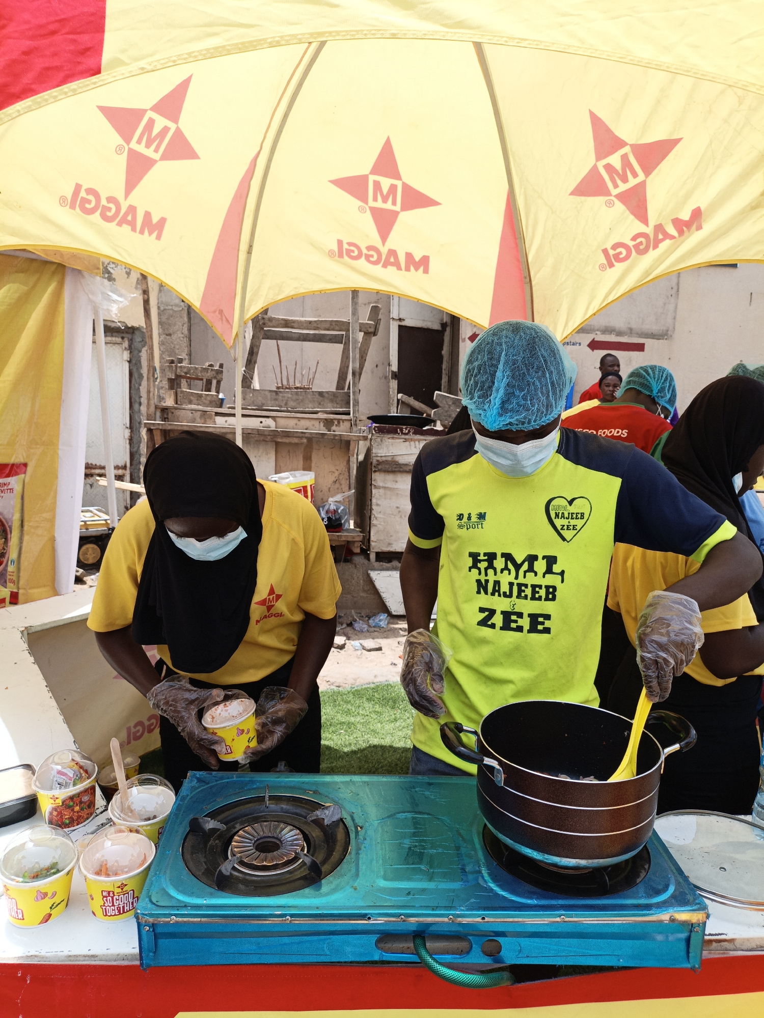 This image captures a street food stall in Tarauni, Nigeria, under a bright yellow umbrella advertising "Maggi." Two individuals, dressed in bright yellow t-shirts and wearing hairnets and face masks, are actively preparing food. One person, wearing a black hijab and a yellow t-shirt, is holding a cup of what appears to be a food product, possibly noodles or soup, while the other, in a neon yellow and navy t-shirt, uses a bright yellow spoon to stir something in a black pot on a teal gas stove.
Several small cups with clear lids are lined up on the counter, some containing food items like vegetables and what looks like a red curry. The text on the cups includes "SO GOOD TOGETHER" and "Maggi." The background shows a somewhat rustic setting with wooden structures and glimpses of other people, some also wearing hairnets, suggesting a communal or event-based food service. The sunlight indicates it's daytime, and the general atmosphere appears busy and focused on food preparation and service. The "Maggi" branding is prominent on the umbrella, suggesting this is a promotional event or a vendor associated with the brand.