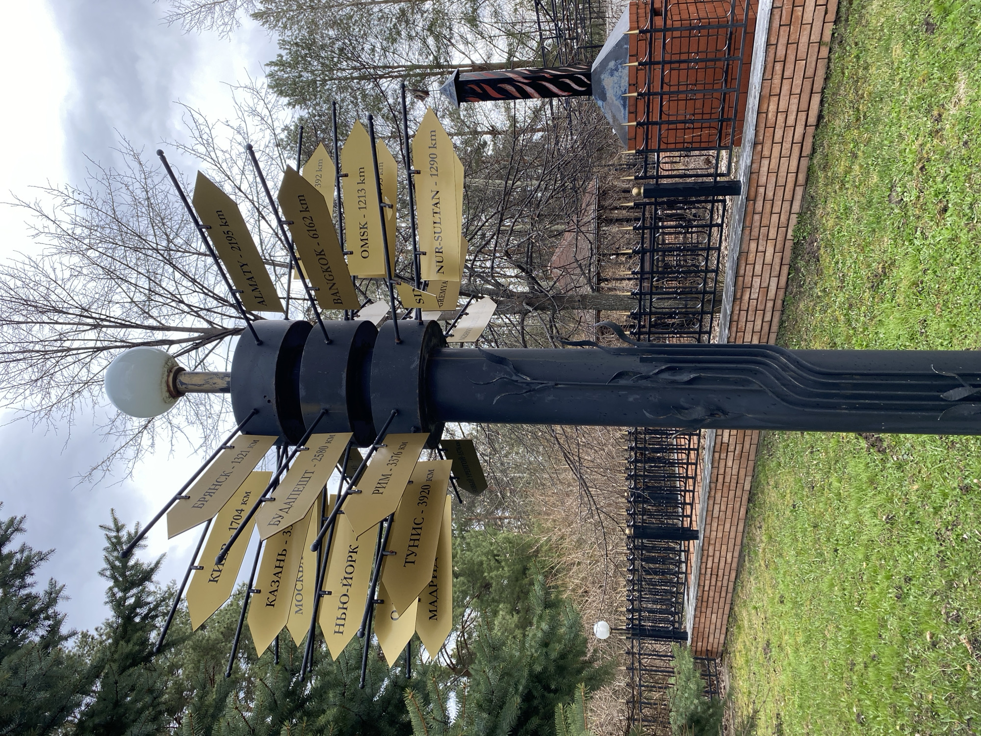 The image displays an outdoor scene in Chaikovsky, Russia, featuring a black metallic signpost adorned with numerous golden, arrow-shaped directional signs. No people are present.
The main subject is the tall, dark signpost, which has a spherical light fixture at its apex. From its central column, multiple signs extend outwards, each pointing in a different direction and indicating a specific city along with its distance in kilometers. The visible text on these signs includes: BRYANSK - 1321 km, KYIV - 1704 km, BUDAPEST - 2580 km, ROME - 3376 km, TUNIS - 3920 km, ALMATY - 2195 km, BANGKOK - 6162 km, OMSK - 1213 km, and NUR-SULTAN - 1290 km. Other partially visible signs include KAZAN, MOSCOW, NEW YORK, MADRID, URENGOY (РЕНГОЙ - 1576 km), a sign with "392 km" and an obscured city, and another sign labeled "YAMAL."
The scene is set in what appears to be a park or public area. The ground is covered with green, though somewhat sparse, grass. In the background, there's a low brick wall topped with a black metal fence, possibly indicating a boundary or an adjacent structure. Deciduous trees with bare branches are visible throughout the background, interspersed with some evergreen trees, suggesting the season is late autumn, winter, or early spring. The sky is overcast, providing soft, diffused lighting and indicating a cloudy daytime. The overall impression is of a landmark or symbolic monument marking a central point with global connections.