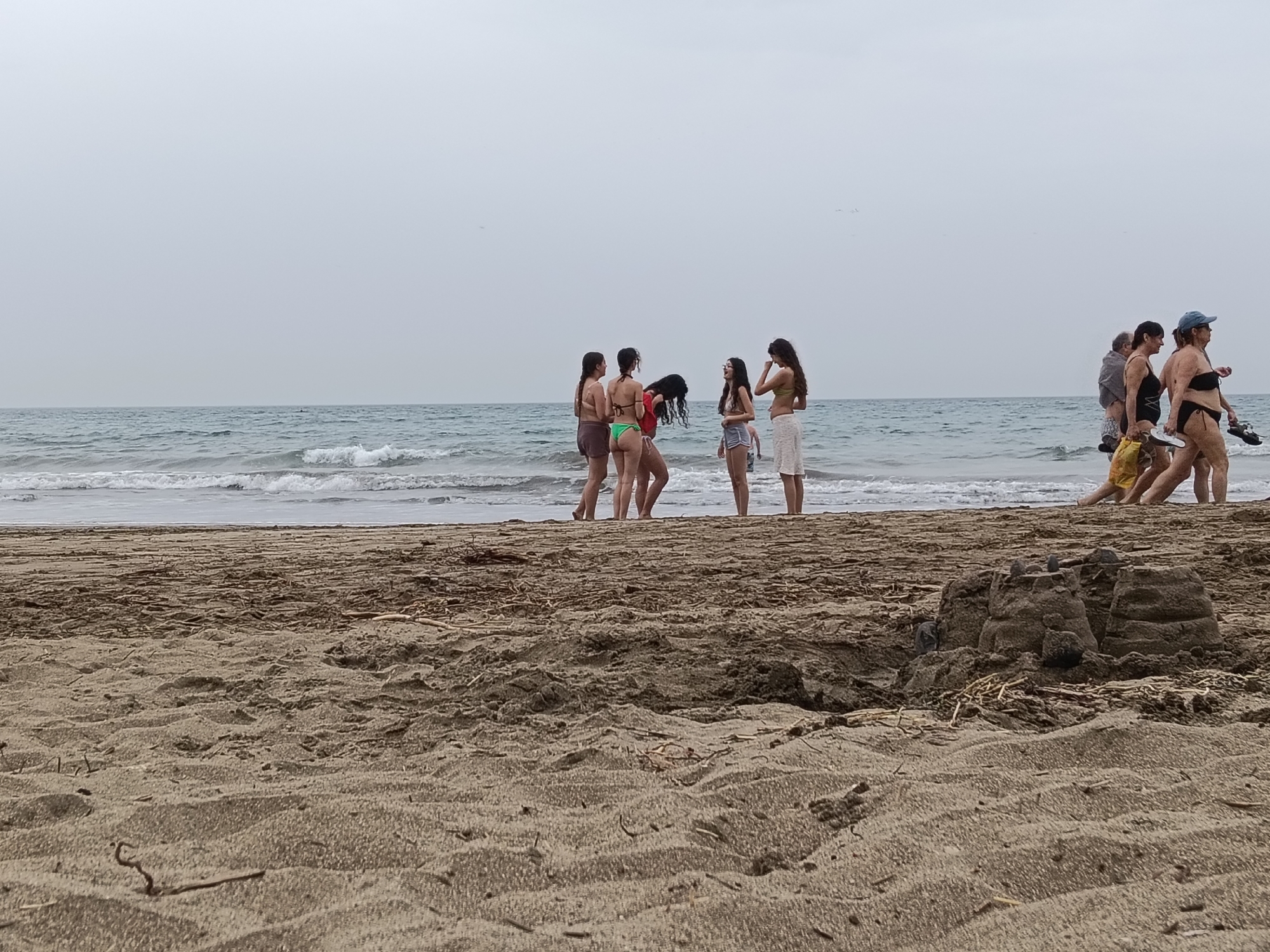 The image captures a serene, overcast day at a sandy beach in San Bartolomé de Tirajana, Spain. The scene is dominated by a wide expanse of light brown sand in the foreground, showing textured patterns and scattered dried organic debris. On the right side of the foreground, a partially eroded sandcastle structure is visible.
In the midground, near the water's edge, two distinct groups of people are present. To the left, a group of five young women or teenagers are standing together, seemingly engaged in conversation. Most are dressed in swimwear, including bikinis and shorts, with some wearing light cover-ups. Their posture suggests a relaxed social interaction. To the right, further along the shoreline, a group of four adults are walking from left to right. They are also dressed in beach attire, with some carrying shoes or bags, indicative of a stroll along the shore.
The background features the calm, blue-grey waters of the ocean with gentle waves breaking near the shore. Above the ocean, a uniformly light grey, overcast sky extends, suggesting a cloudy day without direct sunlight. There is no visible text in the image. The overall impression is one of a casual, leisurely beach day, perhaps during off-peak hours or on a cooler, cloudier day.