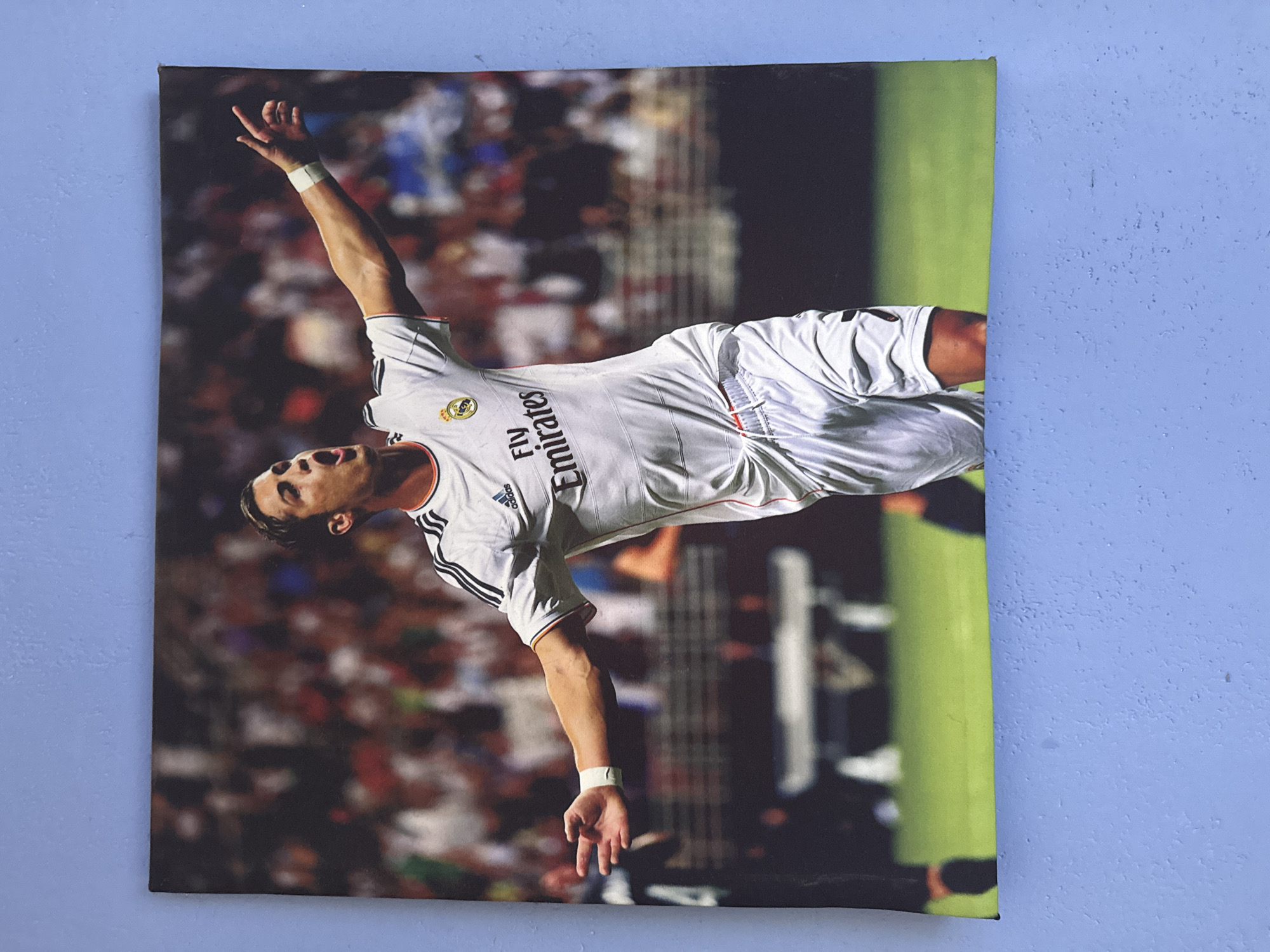 A framed canvas print featuring a portrait of a male soccer player is mounted on a textured, light blue wall. The player is wearing a white Real Madrid jersey with black stripes on the sleeves, white shorts, and a white wristband. His mouth is open in a shout or celebration, and his right arm is raised with his index finger pointing upwards. The background of the print shows a blurred stadium crowd and green soccer field. The print is oriented horizontally. The wall has an uneven, stucco-like texture with visible paint chips.