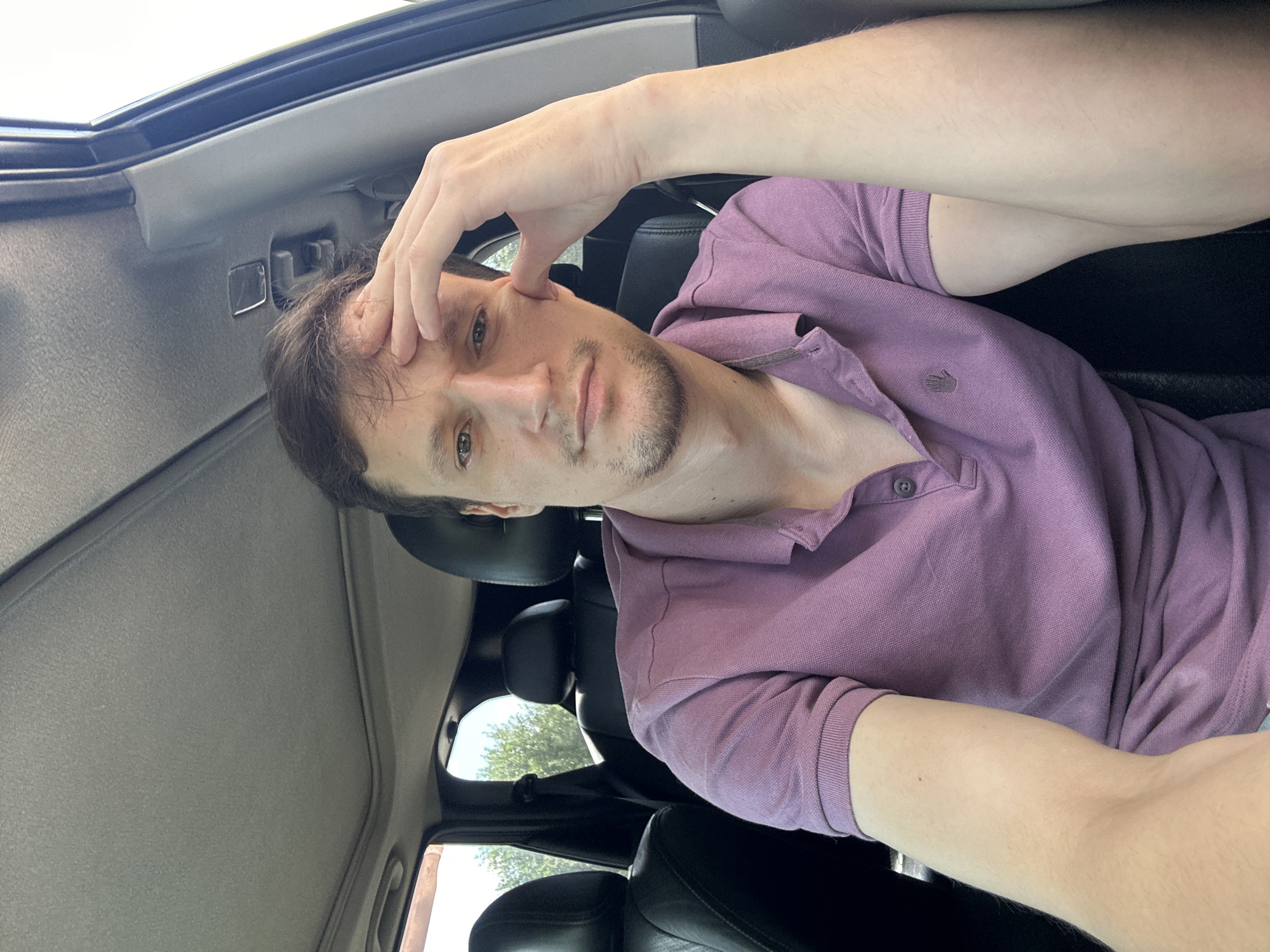 The image shows a man with light brown hair and a beard, wearing a purple polo shirt, sitting in the driver's seat of a car. He has his right hand on his forehead, with his elbow resting on the steering wheel. The car has black leather seats and a sunroof. Outside the car, trees and a blue sky are visible. The image appears to have been taken during the daytime. The location is not specified in the image.