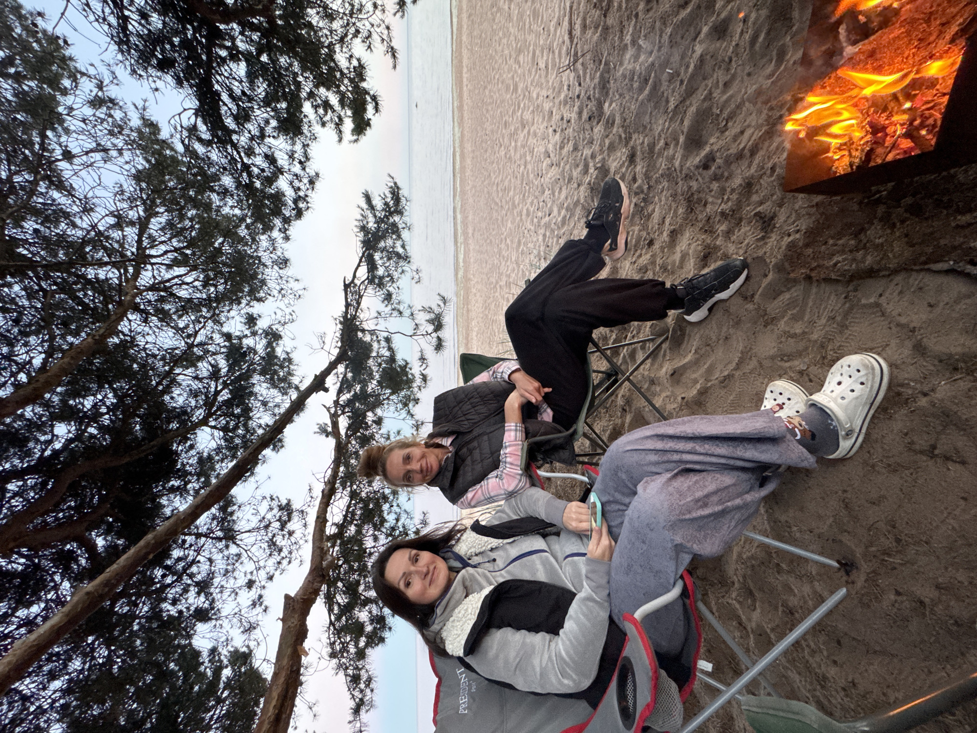 Two women are seated in camping chairs on a sandy beach, facing the viewer. They are positioned under the branches of a pine tree, with a calm body of water and the horizon visible behind them. A campfire burns in a metal fire pit in the foreground to the right. The woman on the left wears a grey hooded jacket, a black vest, and grey sweatpants. She holds a mobile phone in her hand. The woman on the right wears a pink plaid shirt and a black vest, with black pants. Both women appear relaxed. The scene suggests an outdoor gathering on a beach, possibly during dusk. The overall environment is natural, with sand, water, and trees.
