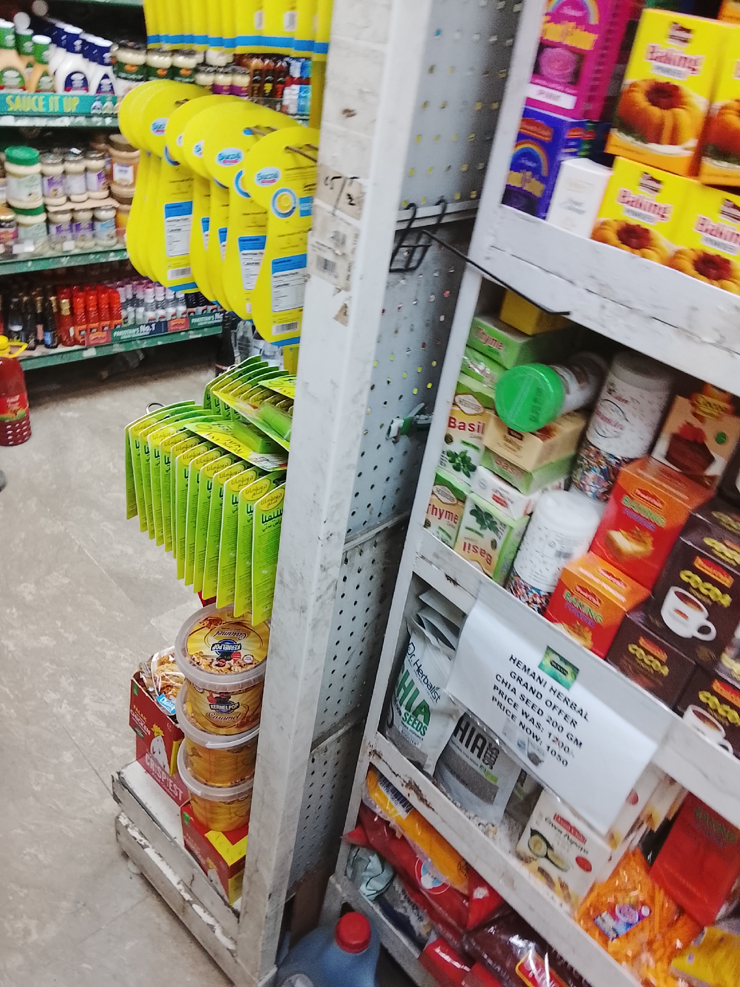 The image shows a shelf in what appears to be a grocery store in Gulgasht Colony, Pakistan. On the left are shelves stocked with sauces, yellow packages of Sucral sweetener, and green packets labeled in Arabic script. Below those are buckets of "Kernel Pop Carmel" popcorn. To the right is a display shelf featuring various products, including baking mixes, spices like basil and thyme, and cocoa. A sign advertises "Hemani Herbal Chia Seed" on sale, with the original price of 1200 reduced to 1050. The setting is indoors, with a brightly lit environment typical of retail spaces. The focus is on the variety of grocery items available.