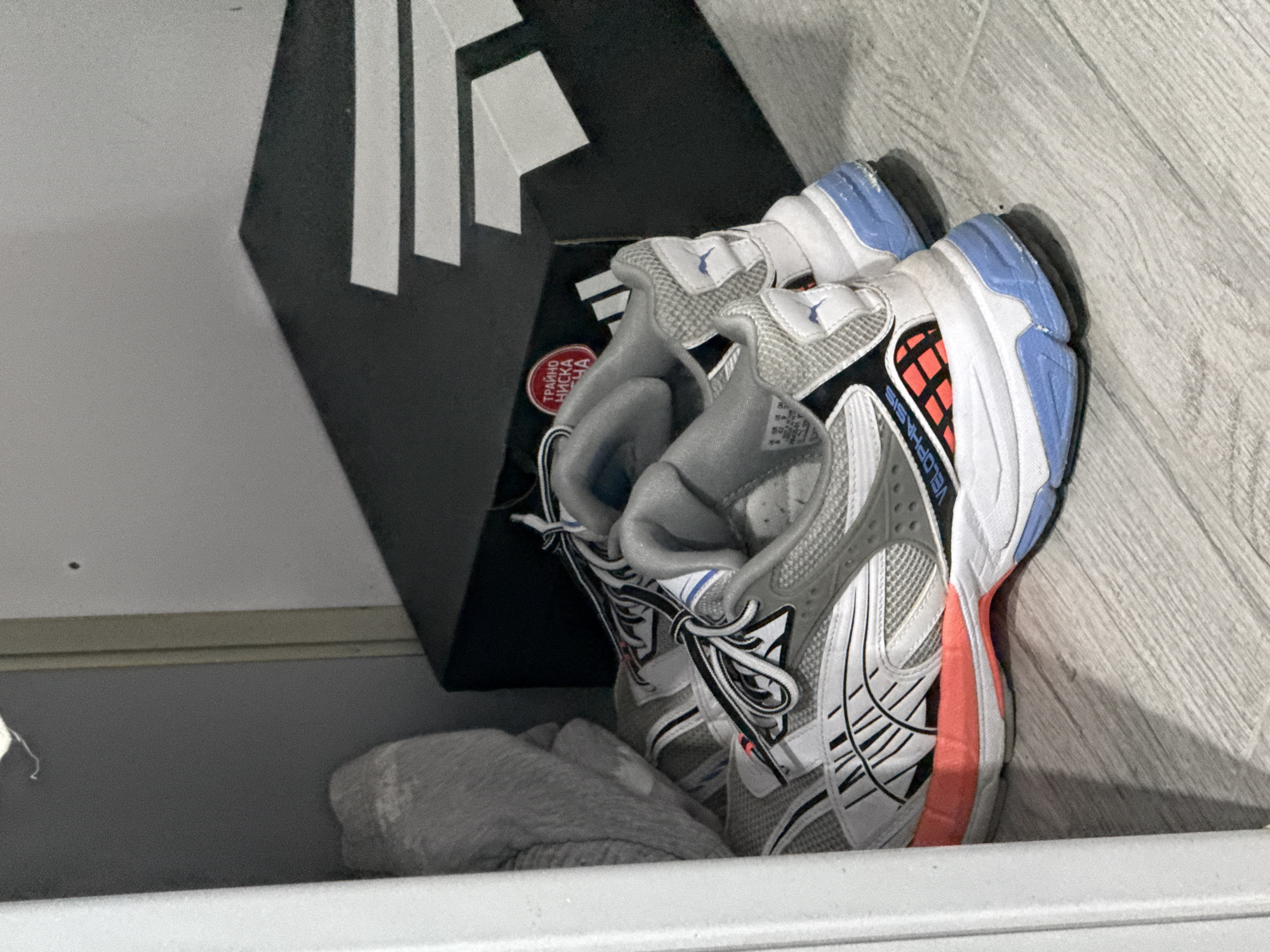 A pair of athletic shoes is prominently displayed, leaning against a dark shoe box and a light-colored wall or floor. The shoes are primarily white and grey, featuring a textured mesh and layered synthetic overlays. Distinctive color accents include light blue on the heel, a vibrant coral or orange section on the mid-sole, and black detailing along the sides. A small blue, bird-like symbol is visible on the tongue of one shoe, while the partial text "ELOPHASIS" or "VELOPHASIS" can be seen on its side.
The shoe box is black with a bold white geometric logo, possibly three vertical bars, positioned on its side. A red circular sticker on the box displays Cyrillic text: "ТРАЙНО НИСКА ЦЕНА," which translates to "LASTING LOW PRICE" or "PERMANENTLY LOW PRICE."
The shoes and box rest on a light, neutral-toned surface, possibly a wooden or laminate floor with a subtle grain pattern. To the left of the shoes, a crumpled grey textile item, resembling a sock or small cloth, is visible. A white horizontal ledge or shelf runs across the bottom of the frame, suggesting an indoor setting such as a closet or storage area. No people or activities are depicted; the image captures a static arrangement of these items.
