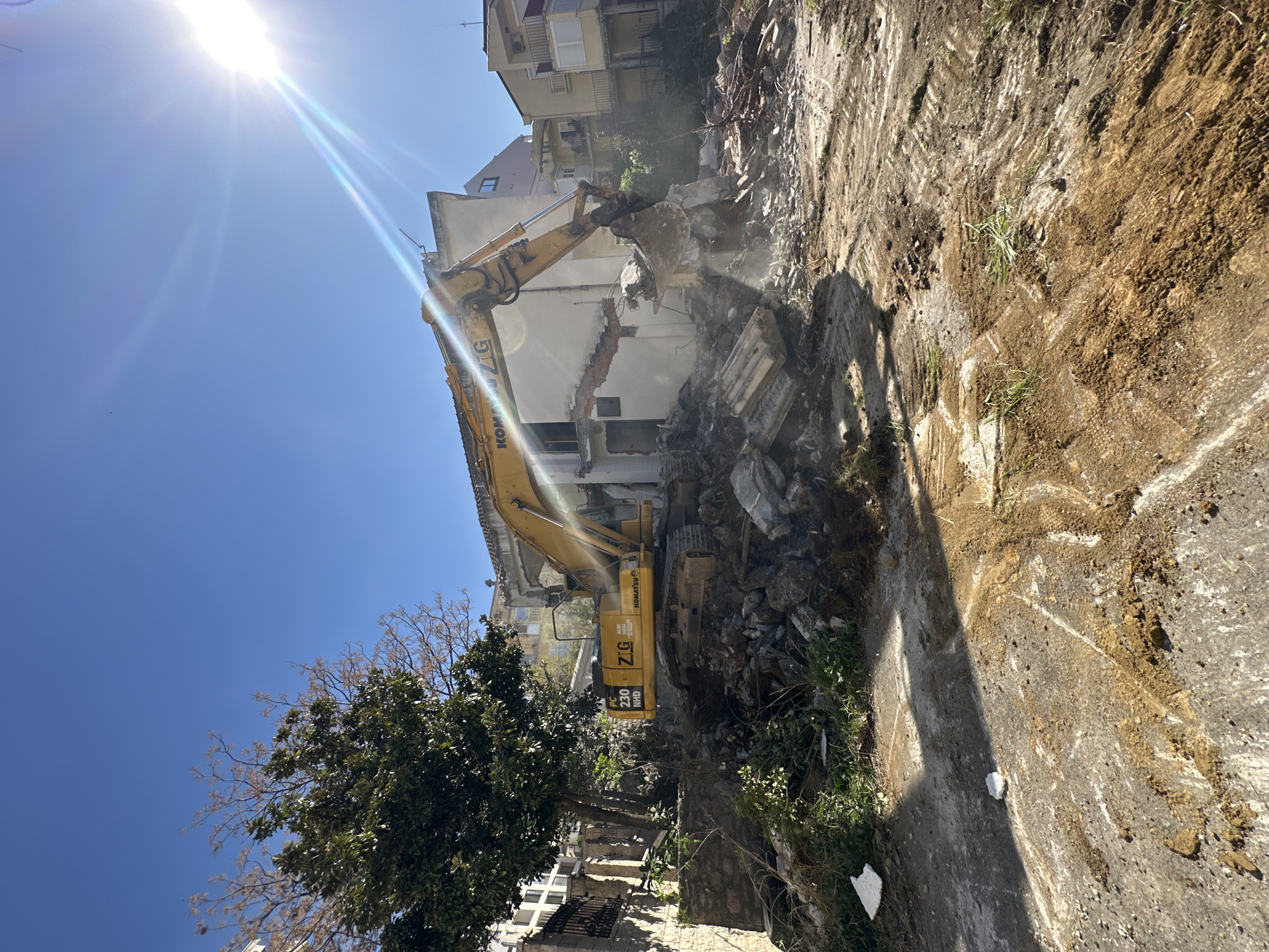 The scene depicts a construction site in Boreti, Montenegro, with the demolition of a building underway. A yellow Komatsu PC 230 NHD excavator is actively tearing down the structure, evidenced by the debris and dust cloud surrounding the machine. The remains of the building consist of concrete and brick, partially dismantled. The site is adjacent to a paved surface and a small patch of vegetation. In the background, multi-story residential buildings are visible. The sky is clear and blue, with a bright sun casting long shadows, indicating a daytime setting. A tree with dense foliage is present near the excavator.
