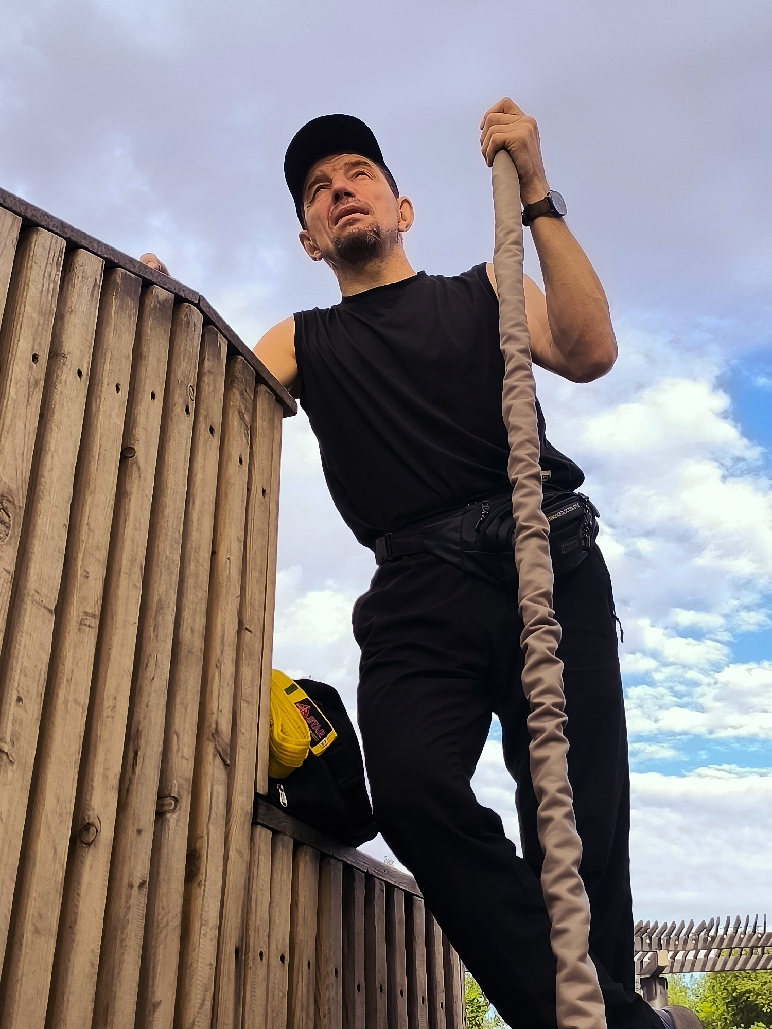 An outdoor, low-angle, medium shot captures a man in black athletic wear, including a sleeveless shirt, pants, and a baseball cap, gripping a thick, beige rope. He is positioned on a wooden structure, possibly a climbing wall or obstacle course, with a blue sky filled with white clouds serving as the backdrop. To his left, a black backpack with a yellow strap is visible, resting on the wooden structure. The man's facial expression is one of exertion or contemplation. The scene suggests an athletic or recreational activity.