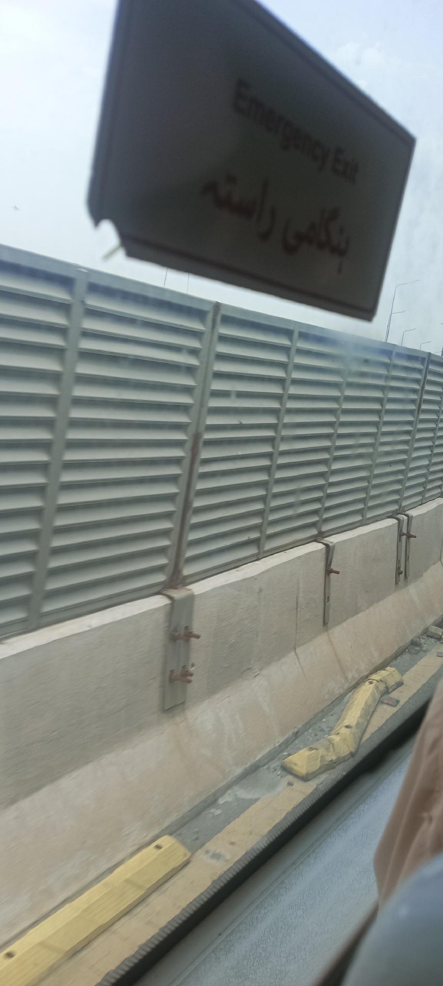 This image shows a tilted view of a highway or main road in Alam Khan Kalay, Pakistan. A reflective sign, partially obscured and tilted, is visible in the upper portion of the frame, displaying text in both English ("Emergency Exit") and what appears to be Urdu script. Below the sign is a gray, slatted sound barrier. The barrier is supported by concrete structures with visible metal brackets and bolts. The foreground shows a portion of the road or pathway with yellow-topped wooden planks and a textured black strip. The lighting suggests it is daytime with an overcast sky.