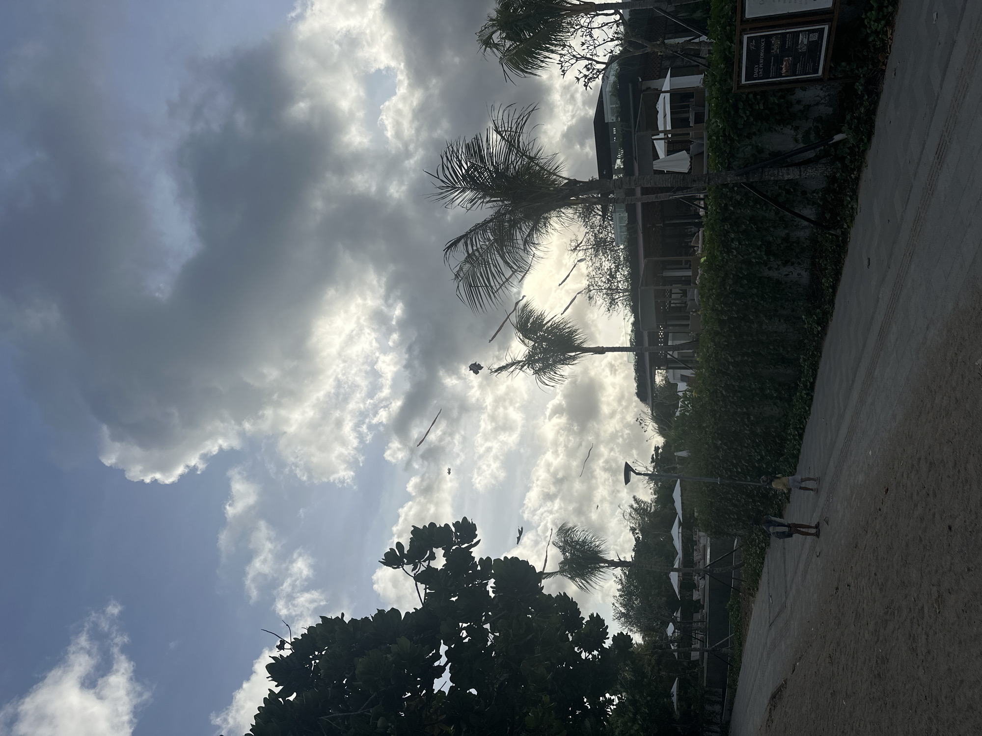 The image captures a scene in Denpasar, Indonesia, featuring a sky with scattered clouds and a mix of sunlight. In the midground, there are palm trees and other foliage. Along the right side of the frame, there is a partially visible building structure covered with greenery. Two individuals are walking on a paved path, and scattered debris or objects are visible in the air, likely indicative of wind.