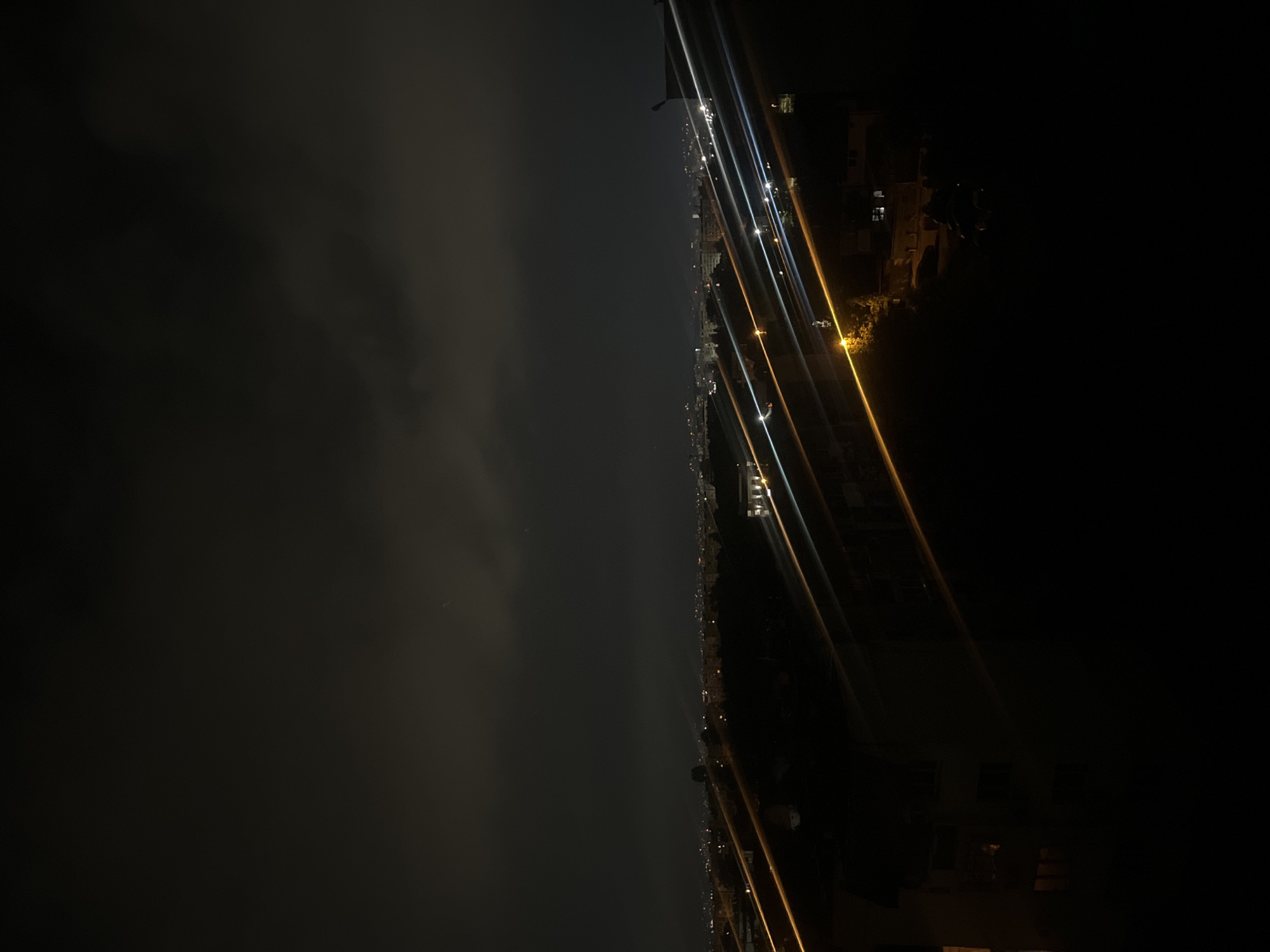 The image depicts a night view of Sofia, Bulgaria, from an elevated position. The city lights stretch out along the horizon, creating streaks and blurs due to the long exposure. Dark clouds occupy a significant portion of the left side of the frame. Buildings are visible on the right, some partially obscured by darkness. Artificial light sources, such as streetlights and car headlights, contribute to the illuminated streaks. The overall environment is dark, with varying degrees of light depending on the distance from the city center.