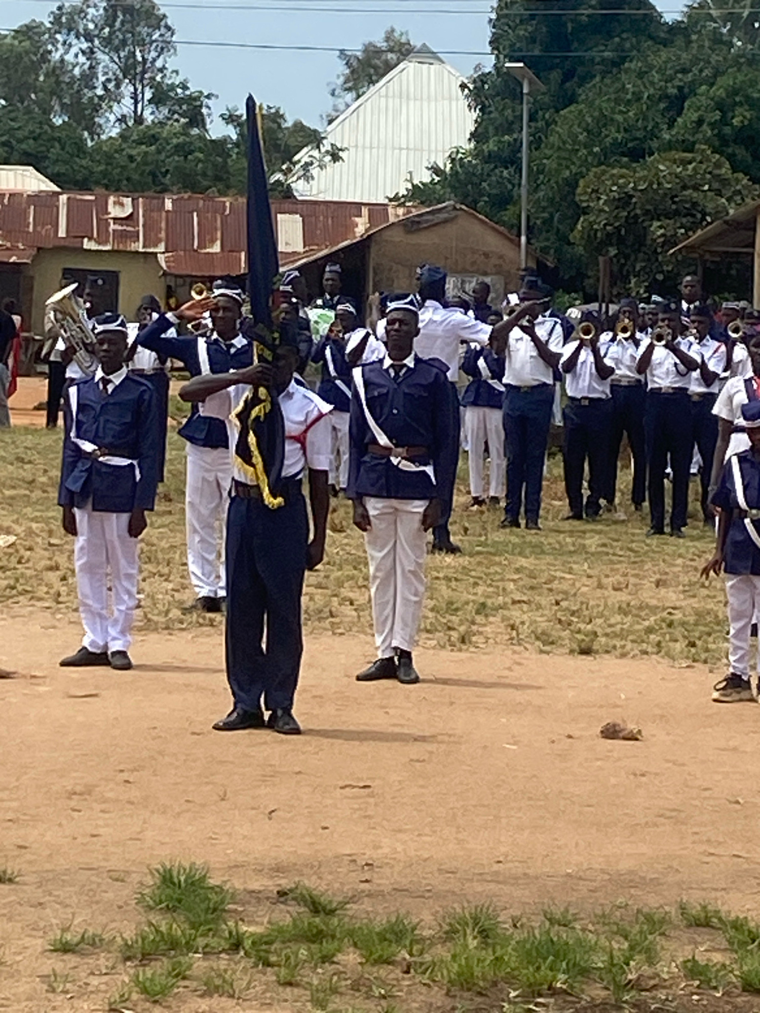In Binchi, Nigeria, a marching band composed of young people in uniform stands in formation on a dirt ground. The individuals are wearing navy blue jackets with white trim, white trousers, and navy blue caps with white bands. Some are holding instruments, including brass instruments like tubas and trumpets, while others are holding a flag. The background features a few buildings with corrugated metal roofs and lush green trees under a bright sky. The scene appears to be an outdoor gathering or parade.