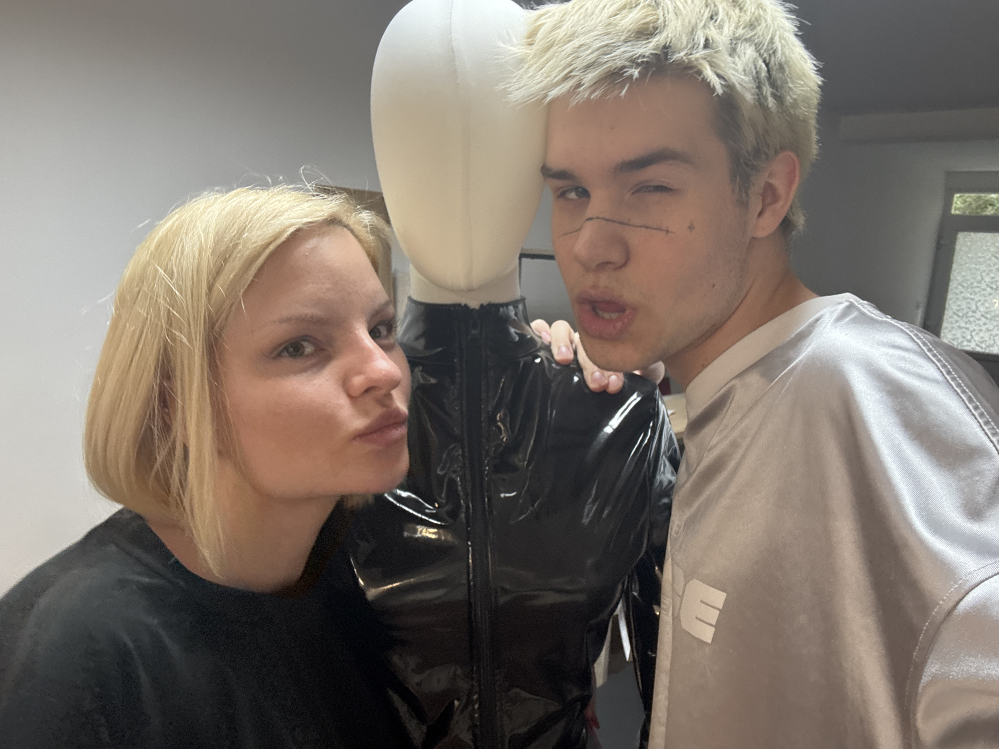 Two people, a blonde woman and a blonde man, are posing with a mannequin in a shiny black jacket. The woman is on the left wearing a black shirt and the man is on the right wearing a gray silky jacket with a white “E” on the right breast. The man has a stitched line drawn on his left cheek. The scene appears to be indoors. A window is visible in the background. There is no information indicating that the location is in the city of Budva, Montenegro.