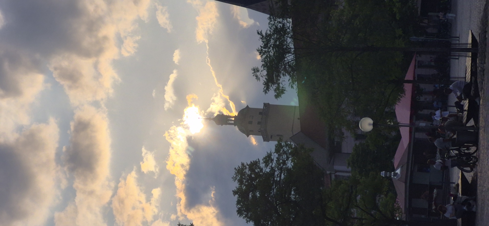 The image depicts a twilight scene in Ingolstadt, Germany, with the iconic Red Tower (Roter Turm) as the central architectural element. The sun is setting, casting a warm glow and creating dramatic light rays that pierce through the clouds, partially obscuring the tower's upper section and its distinctive green dome. The sky is filled with a mix of dark and illuminated clouds, suggesting a dynamic weather condition.
In the foreground, silhouetted by the fading light, are several people gathered in an outdoor seating area. Some are seated on benches, others stand. There are bicycles present, and the area appears to be a public square or plaza, possibly with kiosks or small shops visible behind the seated individuals, indicated by red awnings. Lush green trees frame the lower part of the image, adding a natural element to the urban landscape. The overall atmosphere is peaceful, with individuals engaged in relaxed activities as day transitions into evening.