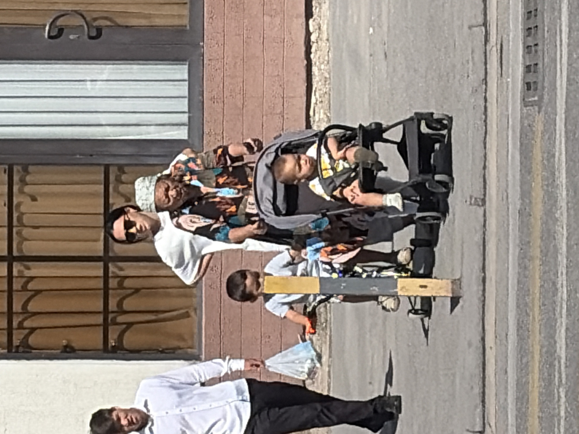 In a sunlit urban environment, a group of people are walking on a paved sidewalk. In the foreground, a young boy sits in a stroller, looking slightly to his left. Beside him, a slightly older child is positioned on a small tricycle, his body turned towards the viewer. Behind these children, an elderly woman with a headscarf is pushing the stroller, wearing a colorful, patterned top. To the left of the children, a man in a white shirt and dark pants walks away from the viewer, carrying a plastic bag in his left hand. In the background, a woman wearing sunglasses stands behind the elderly woman. The scene takes place in front of a building with a textured reddish-brown facade and dark-framed windows covered with vertical blinds or drapes. The pavement is grey and shows some wear. Yellow and white lines mark a street in the lower part of the image.