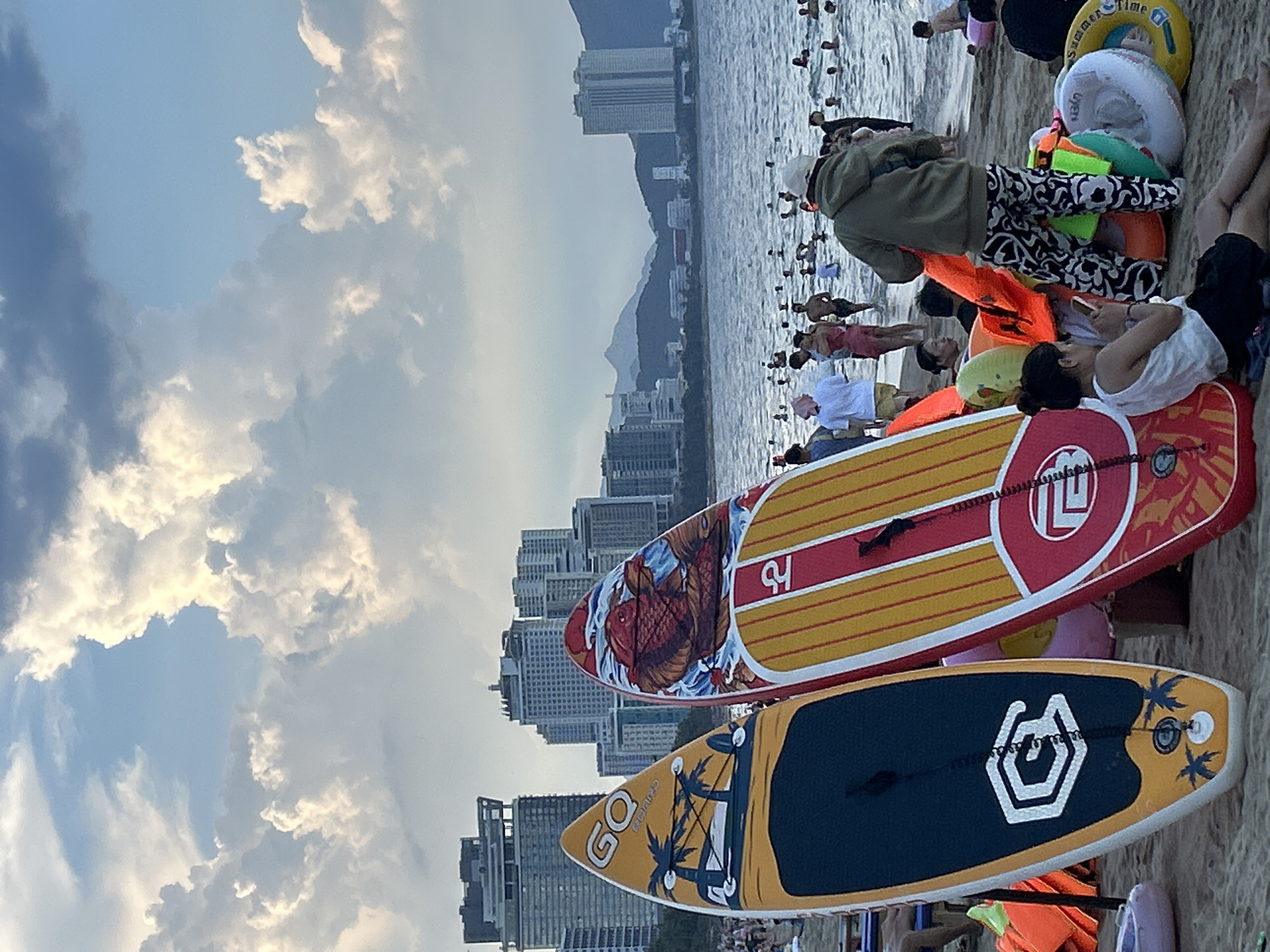 This is a medium shot photograph of a beach in Nha Trang, Vietnam, at sunset. The foreground is dominated by two paddleboards standing upright on the sand, one yellow and blue with palm trees, and the other red and yellow with a koi fish design. In the middle ground, people are wading and swimming in the ocean. The background features a cityscape with tall buildings, mountains, and a cloudy sky. The overall atmosphere is one of leisure and recreation.