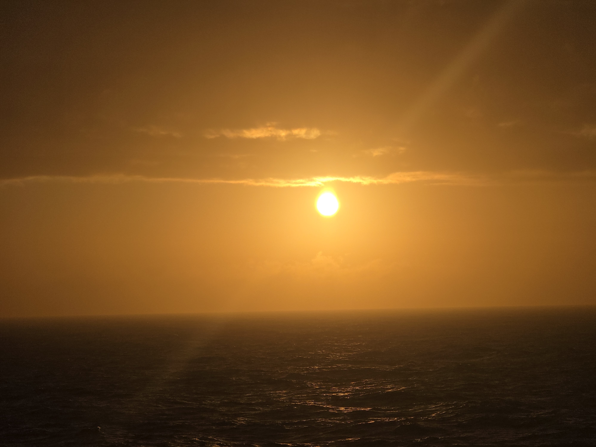 A low-angle, wide shot depicts a horizon dominated by the sun setting (or rising). The intense light of the sun is partially obscured by a thin cloud band running horizontally across the image. Above the cloud bank, the sky is a consistent gradient of golden orange. Below the cloud bank, the glow of the sun diffuses into a hazy horizon line, blending into the dark ocean waves that take up the lower portion of the image. The water is not calm, showing significant surface movement. A single, faint ray of light extends from the top right corner towards the sun. The location is indicated only as "Unknown, Unknown". No people or specific objects are visible in the image.