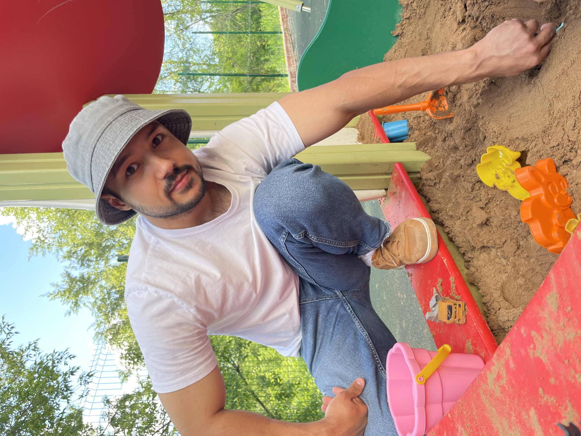 The image captures a man interacting with a sandbox at an outdoor playground in Izhevsk, Russia. The man, with short dark hair, a mustache, and a short beard, wears a light grey bucket hat, a white short-sleeved t-shirt, and blue jeans. He is positioned in a crouch, his right arm reaching into the sand as if playing or digging, while his gaze is directed towards the viewer.
The sandbox is filled with light brown sand and contains various colorful plastic toys, including an orange spade, a blue bucket, yellow and orange animal-shaped sand molds (resembling a chick and a lion/cat), a small yellow toy construction vehicle, and a pink bucket with a yellow handle. Red and green playground structures partially frame the foreground and background. Beyond the playground elements, green trees and a clear blue sky are visible, indicating bright, sunny daytime weather. A white mesh fence or netting can be seen in the distance through the foliage. The camera angle is low, looking up at the man, creating a dynamic perspective of the scene. The man's expression is neutral. No discernible text is visible.