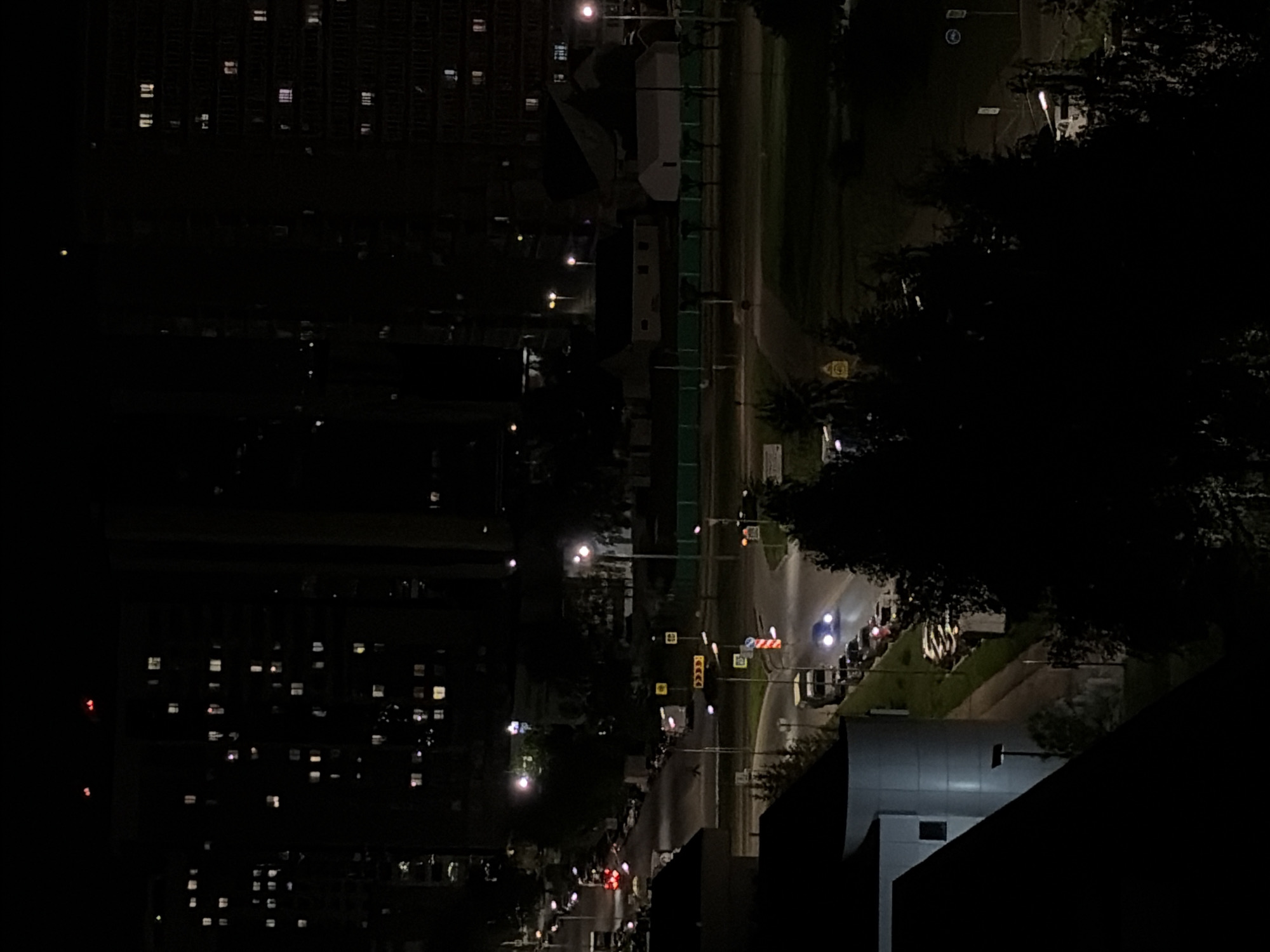 This nighttime shot shows a street scene in Naberezhnye Chelny, Russia. High-rise apartment buildings with illuminated windows dominate the left side of the frame. The right side of the image features a dark street with sporadic streetlights, traffic lights, and a few cars. Trees line the street. The bottom right corner has a light-colored roof, possibly a building's extension.