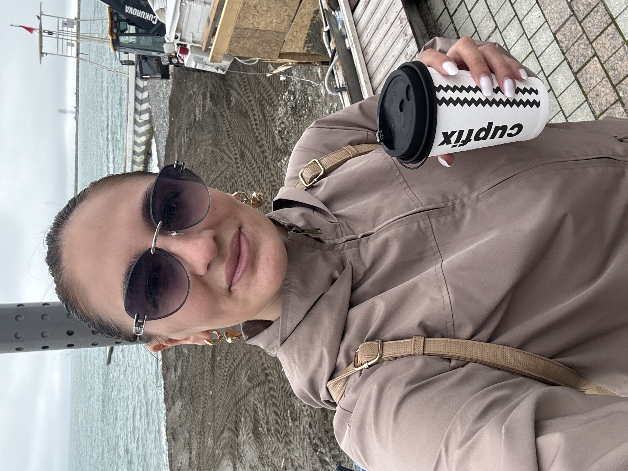 A woman in a beige jacket and sunglasses holds a "cupfix" branded coffee cup with white zigzags and a black lid in her right hand. She is on the coast, with the sea and beach in the background. A construction crane and other structures are also visible behind her. The location appears to be in Sochi, Russia, based on the "CUKUROVA" marking on the crane. The surroundings include metal poles, a dirt beach area with tire tracks, and paved sidewalks. The woman's nails are painted white. She is wearing gold hoop earrings. A beige cross-body bag strap is visible across her jacket.