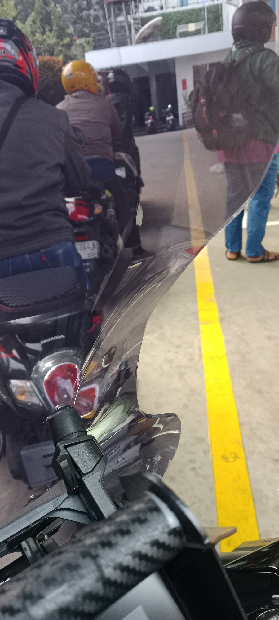 The image shows a group of people on motorcycles, likely in Tangerang, Indonesia. In the foreground, a motorcycle's windshield and handlebars are visible. The windshield is clear and appears to have some scratches or marks on it. The handlebars are black and have a carbon fiber texture. Behind the windshield, a red taillight of a motorcycle is visible, along with the license plate which reads "B 5744 AB".
In the mid-ground, several people are seated on motorcycles. One person in the foreground is wearing a black jacket and blue jeans, with a black and red helmet. Another person behind them is wearing a brown jacket and a yellow helmet. Further back, another person is wearing a dark jacket and a black helmet. The people are facing away from the camera, suggesting they are either waiting or in transit.
The background shows a building with a white facade and some greenery. A yellow line is painted on the ground, indicating a lane or a boundary. The overall environment appears to be an urban setting, possibly a parking area or a street. The presence of multiple motorcycles and people dressed in casual attire suggests a typical day in Tangerang.