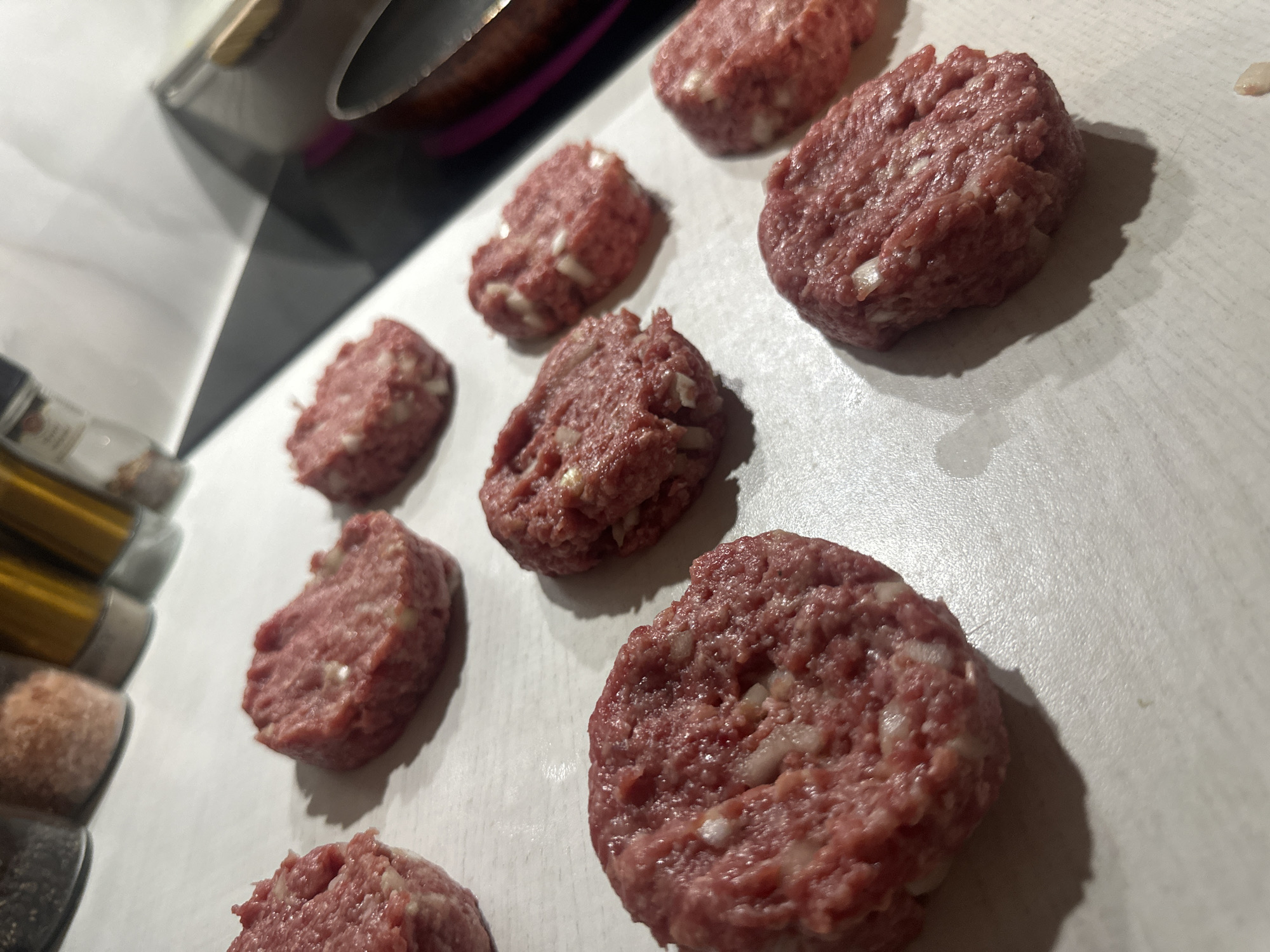 The image displays a close-up, high-angle view of multiple raw meat patties arranged on a light-colored surface, likely a kitchen countertop or large cutting board. There are at least eight distinct patties visible, with their shapes varying slightly, suggesting they were hand-formed. Each patty is a reddish-pink color, characteristic of raw ground meat, and contains small, irregularly shaped white inclusions, which appear to be chopped onions or fat.
In the upper left corner of the frame, the edge of a metallic pot or pan and part of a dark-colored kitchen appliance are visible. Along the far left side, several spice shakers or bottles are stacked vertically, partially visible and out of focus. Any text on their labels is unreadable due to the angle and blur.
The scene suggests the activity of food preparation, with the patties likely assembled for cooking, such as making burgers or meatballs. Shadows cast by the patties indicate an indoor light source from the top-left. The overall setting appears to be a domestic kitchen. No specific time of day, weather, or discernible emotions are conveyed.