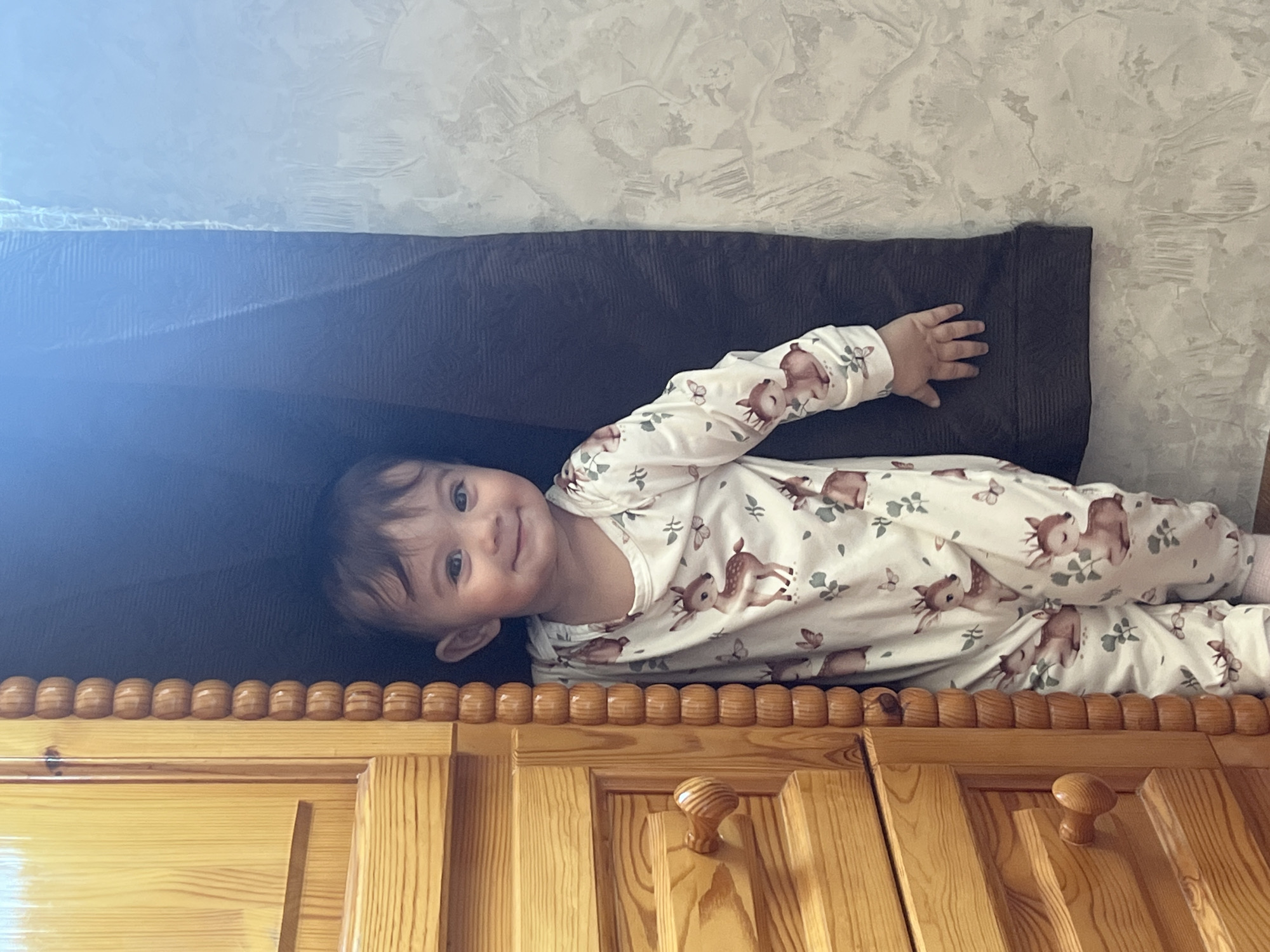 A young child lies on their side, likely on a bed or elevated surface, in a room in Elena, Bulgaria. The child is wearing a white onesie with a pattern of brown deer and butterflies. They are smiling at the camera. Behind the child is a dark brown, corduroy-like material. Below the child is a wooden structure, possibly a cabinet or headboard, featuring decorative wooden knobs and vertical wood panels. The background wall is textured and appears to be painted a light color.