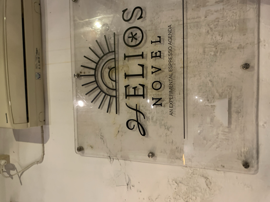 The image shows a clear acrylic sign mounted to a white wall. The sign is attached by silver screws. The sign reads "HELIOS NOVEL, AN EXPERIMENTAL ESPRESSO AGENDA" with an image of a sun above the text. The wall has some damaged areas and is dirty. An air conditioner is mounted above the sign. The coordinates -7.751, 113.213 place this location in Probolinggo, Indonesia.