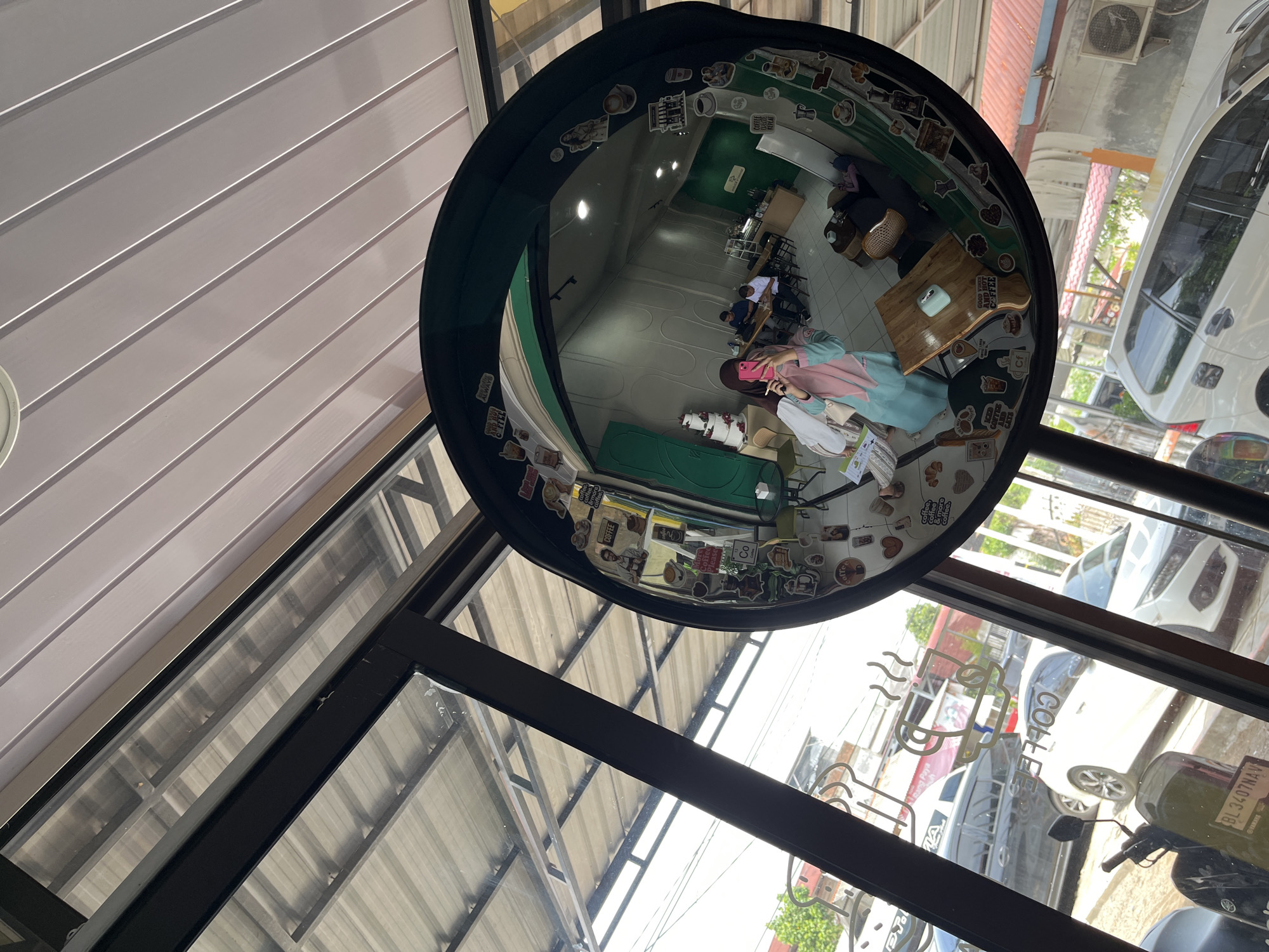 The image shows a reflection in a large convex mirror, likely located inside a cafe in Lhokseumawe, Indonesia. The mirror is adorned with various stickers, many of which feature coffee-related imagery and text.
Inside the cafe, visible in the reflection, a woman wearing a hijab and a turquoise top is taking a selfie with a pink phone. She is standing near the entrance, which appears to be a glass door or window. Further inside the cafe, two men are seated at a table, one with a laptop. There are other tables and chairs, a counter area, and a tall cake displayed on a table. The walls are painted a light gray with a textured design, and a section of the back wall is painted green.
Outside the cafe, through the glass, a street scene is visible, including parked cars and a motorcycle. The presence of "BL 3407 NA" on the motorcycle's license plate provides a clue to its Indonesian origin. The architecture and vegetation suggest a tropical or sub-tropical climate. The overall impression is that of a casual, modern cafe setting.