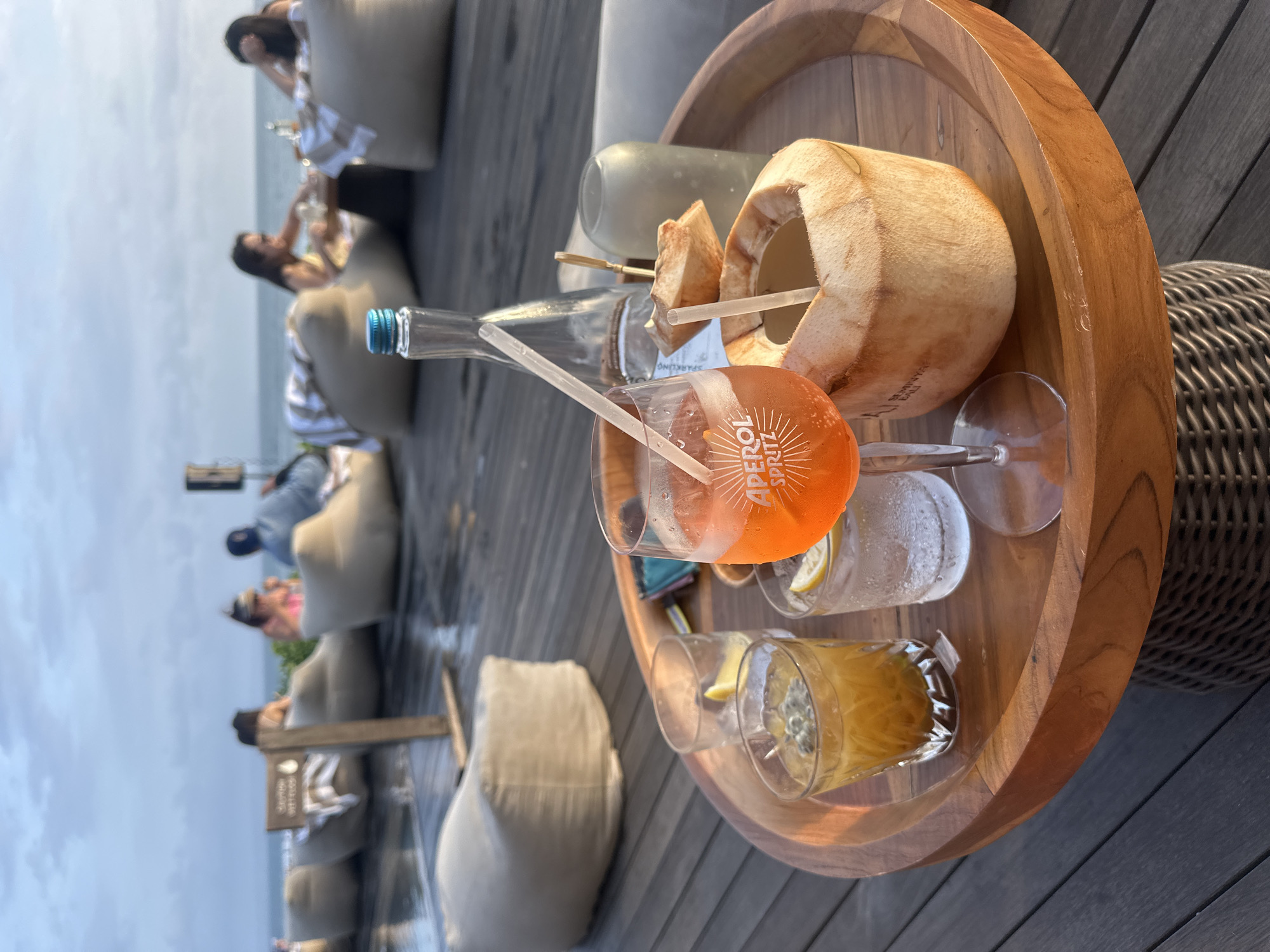 A wooden tray holds several drinks and a whole coconut with a straw. In the foreground, an orange Aperol Spritz is in a stemmed glass with a white straw. To its left is a clear glass with ice and a yellow drink, possibly passionfruit juice, topped with seeds. Next to that is a glass of clear liquid with ice and a slice of lemon. To the right of the Spritz is the halved coconut with a straw in it. Behind the drinks are a glass bottle with a blue cap, a short skewer with pieces of coconut, and a frosted glass. In the background, people sit on large, beige beanbag chairs facing the ocean. The scene appears to be on a wooden deck at dusk.