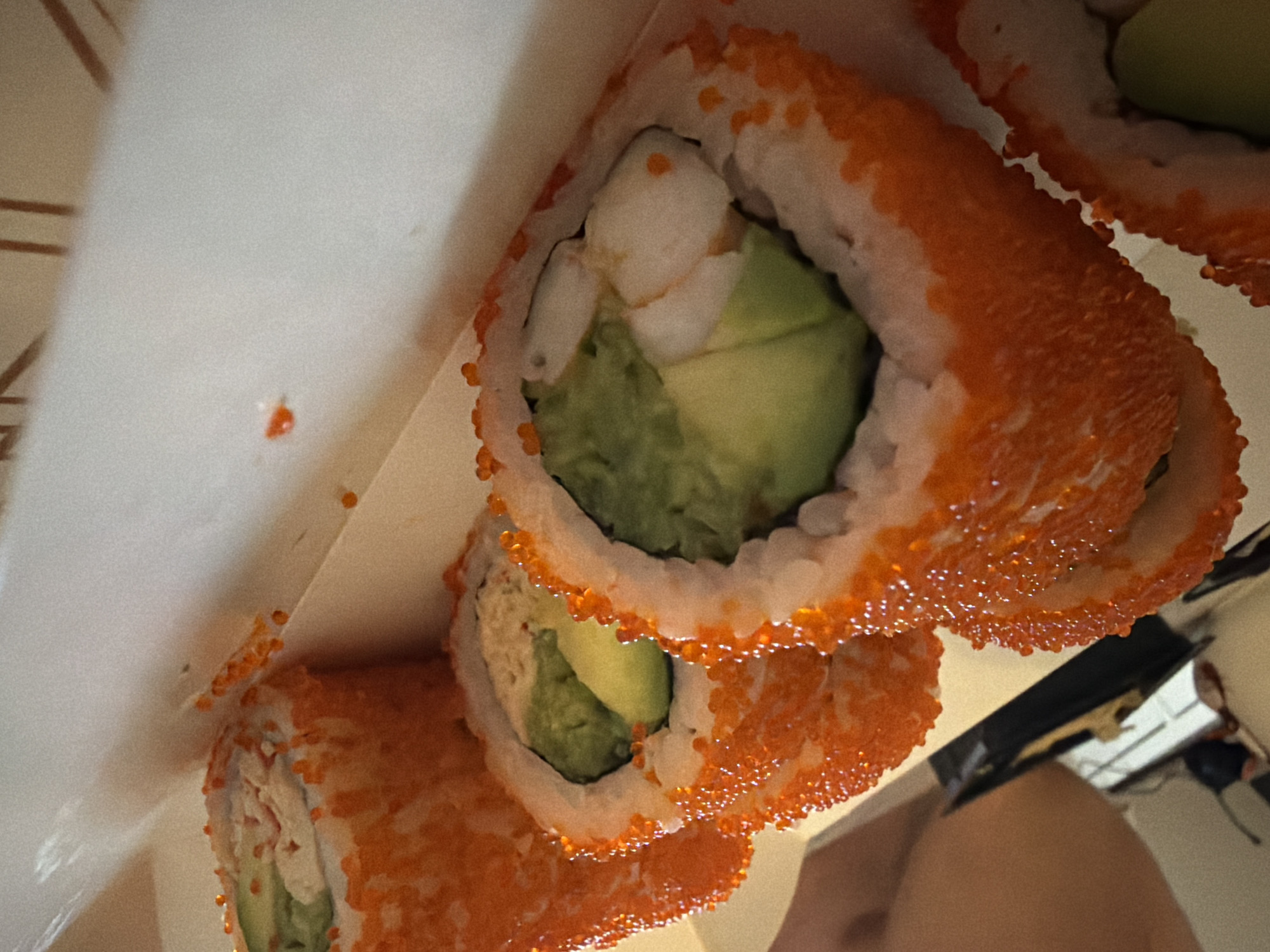 The image shows a close-up of several pieces of sushi, likely a California roll, arranged in a white container. The sushi consists of white rice, avocado, and possibly crab stick, and is coated with orange fish roe (tobiko). The background includes portions of what appears to be a home interior, with hints of furniture and electrical outlets. It is impossible to determine if this photo was taken in Budva, Montenegro from the image alone.