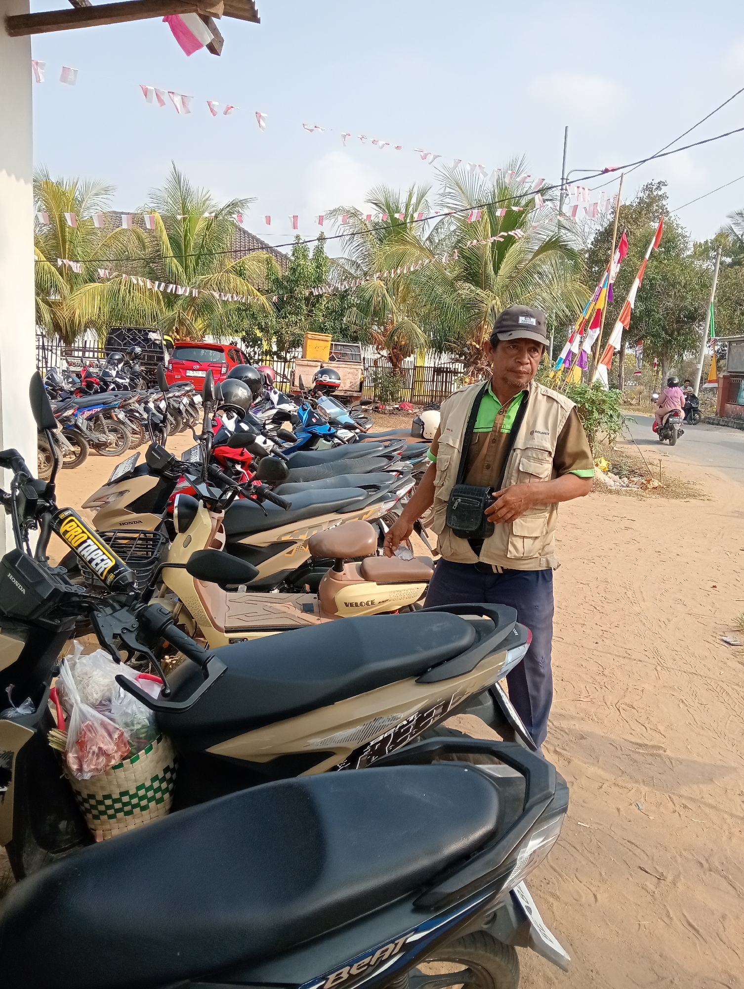 In Gunung Sugih, Indonesia, a man stands beside a row of parked motorcycles under a clear sky. He is wearing a beige vest over a green shirt and dark pants, with a black cap on his head and a black bag slung across his chest. Several motorcycles are parked in the foreground and background, some with helmets on the handlebars. In the background, there is a red car, a building with a white pillar, and trees with Indonesian flags (red and white banners) strung between them. A dirt road is visible to the right with a person riding a motorcycle. The scene appears to be an outdoor parking area.