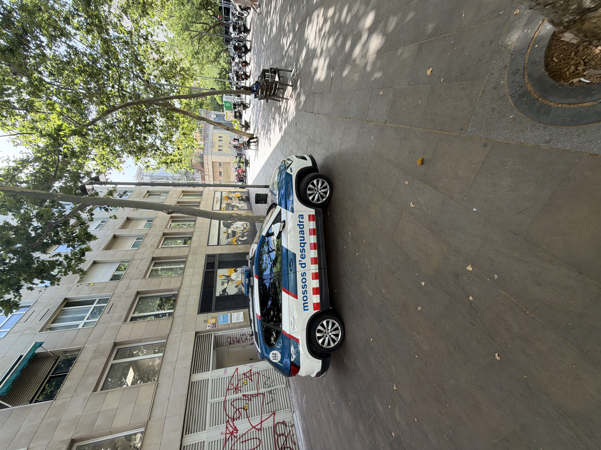 The image depicts a police car, identified as belonging to the "Mossos d'Esquadra," parked on a sidewalk in Barcelona, Spain. The car, a white SUV with blue and red markings, has its emergency lights illuminated on the roof. The vehicle is positioned on a tiled sidewalk in front of a modern building with a light-colored facade and several windows. The building appears to be commercial or residential. To the right of the police car, trees with green foliage provide shade, and a street with parked motorcycles is visible. A few individuals are seen in the background, with one person sitting on a bench. The overall environment suggests a daytime scene in an urban setting.