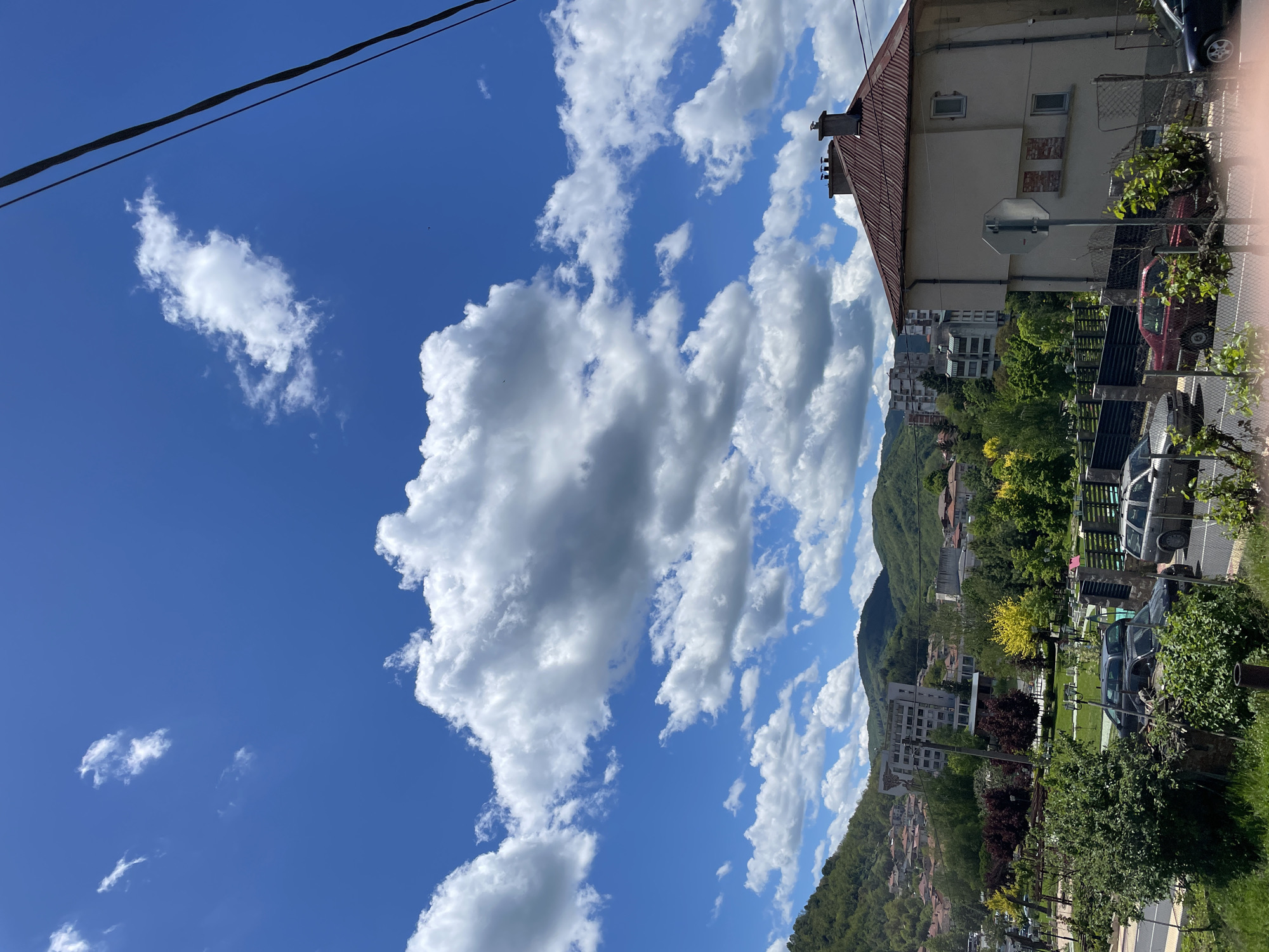 A street scene captured in Zlatograd, Bulgaria, features a building with a brown roof and light-colored walls, partially visible on the right side of the frame. A mountain covered with trees is visible behind the building and down below other buildings. Several cars are parked on the street along a fence. A patch of grass and trees can be seen in the foreground. Overhead powerlines are on the upper left, contrasting with a bright blue sky filled with numerous white clouds.