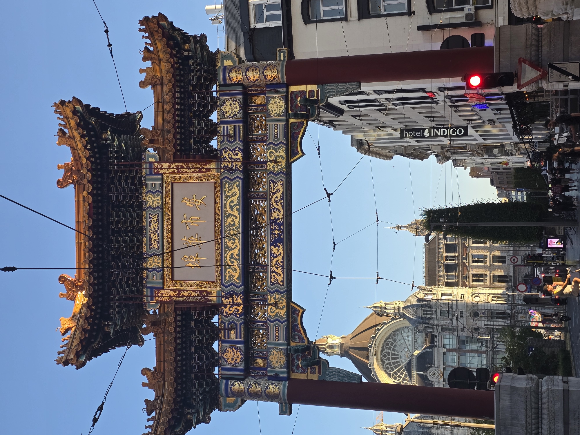 The image captures a view through the ornate Chinese archway in Antwerp's Chinatown, illuminated by warm sunlight against a clear blue sky. The archway, a prominent landmark, features intricate gold and blue detailing, with Chinese characters inscribed on a central plaque. Below the arch, the cityscape unfolds. To the left, a grand building with a domed roof and arched windows, likely Antwerp's central station, is visible. The street is lined with buildings of various architectural styles, some featuring traditional European facades. Traffic lights, street signs, and utility wires crisscross the scene, indicating an urban environment. Several people are seen walking and cycling on the street. A red traffic light is illuminated on the right side of the image. The overall impression is a vibrant street scene in a European city with a significant Chinese cultural presence.