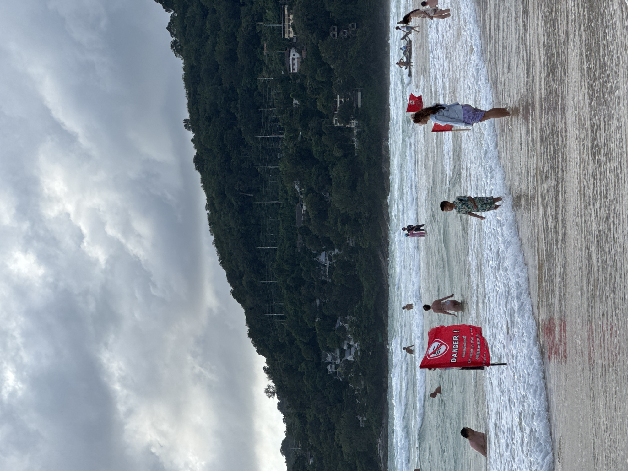 The image captures a view of Karon Beach in Karon, Thailand, under a cloudy sky. In the foreground, a red danger flag with text in Thai and English warns of hazardous conditions. Several people are visible in the shallow waters and on the sandy shore, with some wading and others swimming. The ocean has moderate waves, creating white foam along the shoreline. In the background, a lush, green hillside is covered with trees and dotted with white buildings. The overall scene depicts a typical day at a beach with varying weather conditions and recreational activities.