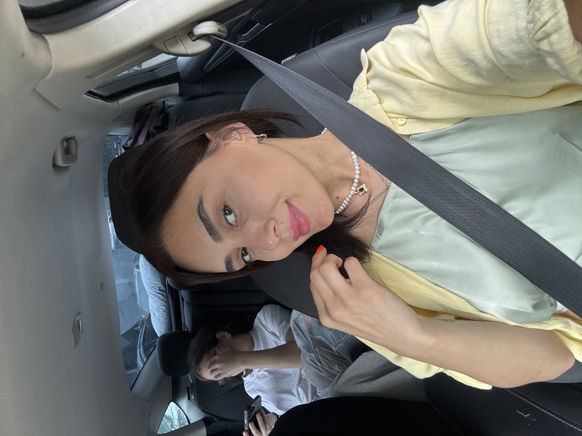 In the foreground, a woman with dark hair is seated in the passenger seat of a car, facing the camera. She is wearing a pale yellow collared shirt over a light green sleeveless top and a pearl necklace with a green pendant. A gray seatbelt crosses her chest. Her right hand is near her face, and her left arm is bent at the elbow, resting on her lap.
In the background, a younger individual with brown hair and glasses is seated in the back seat of the car, facing left. They are wearing a white t-shirt and gray pants and are holding a mobile phone in their left hand, with their right hand covering their face. A light-colored pillow is visible on the back seat behind them. The interior of the car is visible, including the headrests and a portion of the car's ceiling. Outside the car windows, glimpses of other vehicles and what appears to be a somewhat overcast sky can be seen. The image was captured in Tbilisi, Georgia.