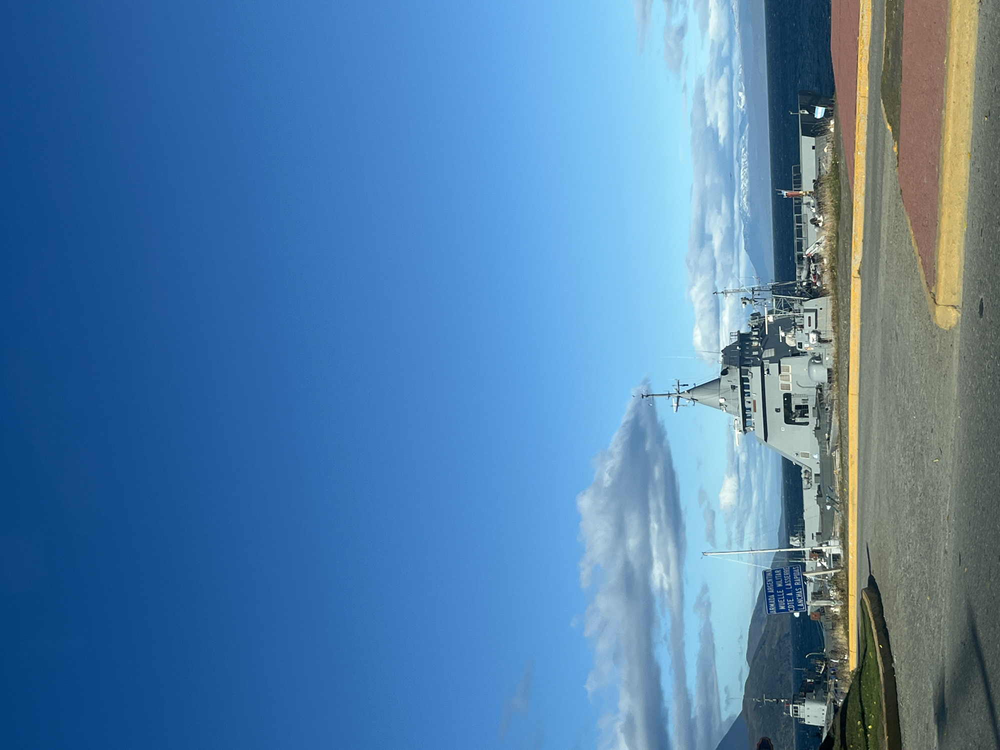 The image depicts a waterfront scene in Ushuaia, Argentina. Several gray naval ships are docked along the pier. A street runs adjacent to the pier, marked with a yellow line. A sign in Spanish indicates "Armada Argentina", "Muelle Militar," and "Lanchas Rapidas". The backdrop includes a blue sky with clouds and mountains.